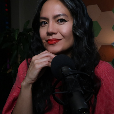 A woman with dark wavy hair, red lipstick, and a nose piercing smiling in front of a microphone, with a geometric pattern wall in the background.