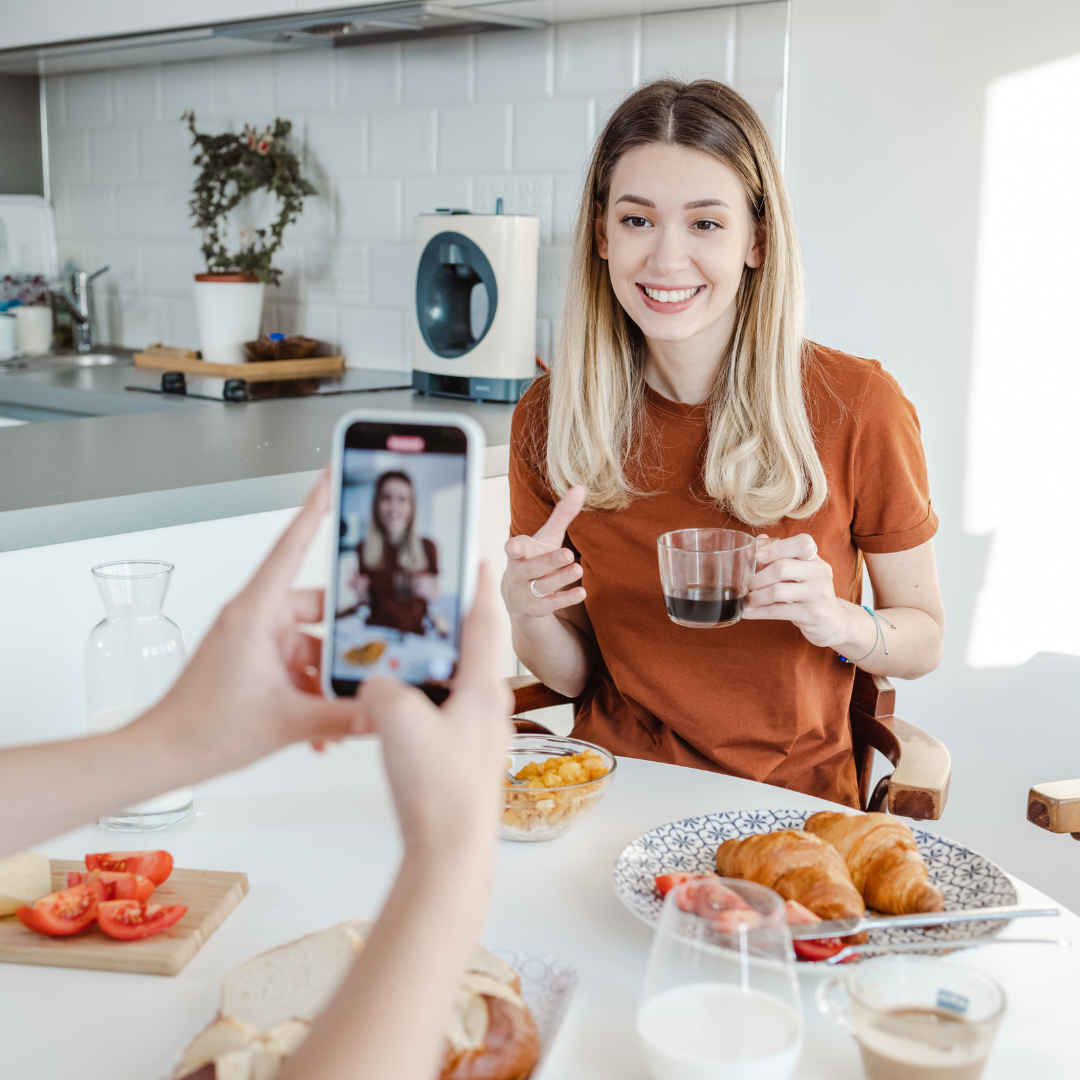 Woman having breakfast at a table, holding a glass of coffee, while being photographed with a smartphone. The table is set with croissants, tomatoes, bread, and a glass of milk.