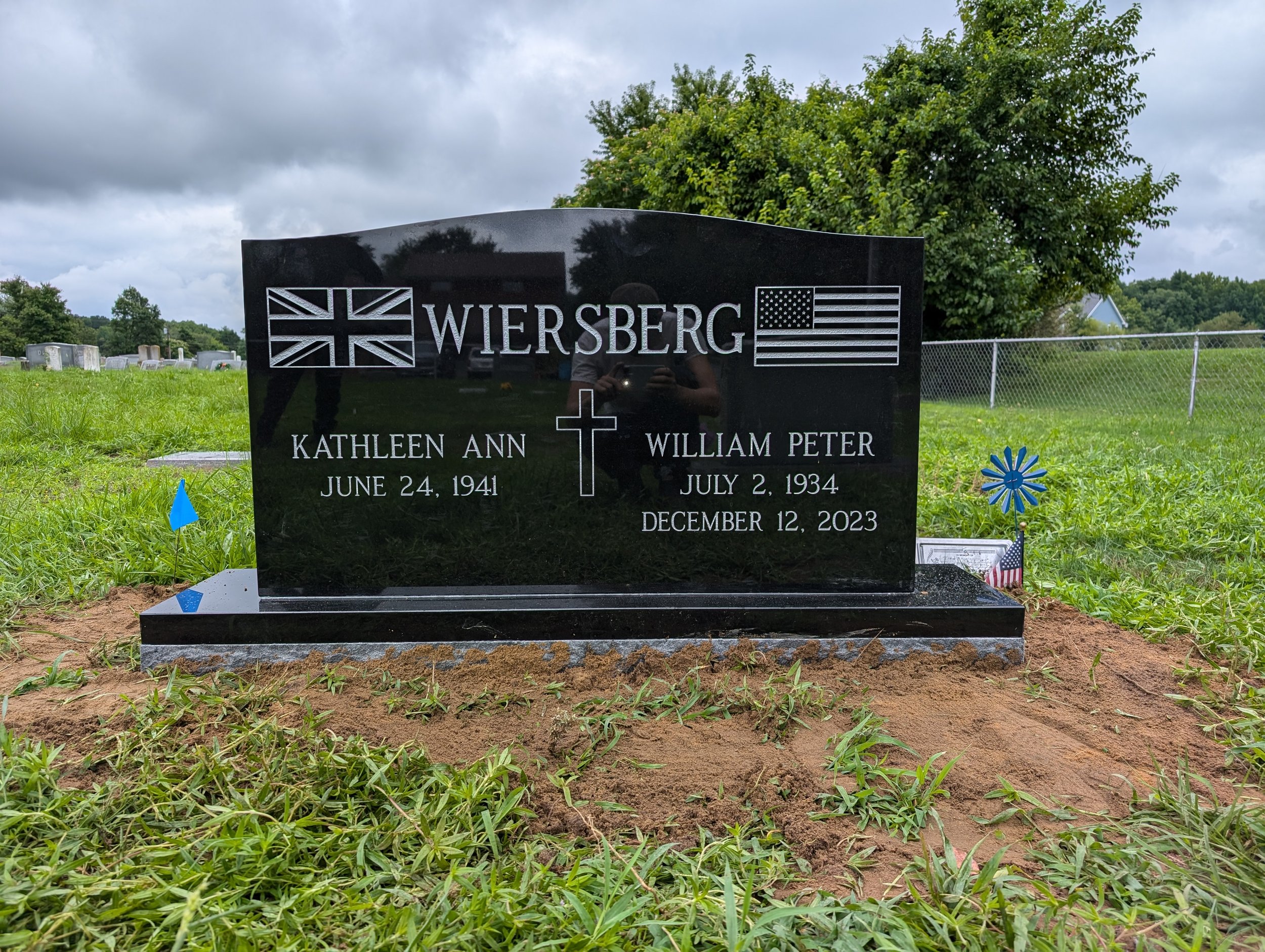 Gravestone with the name 'Wiersberg,' symbols of the British and American flags, a cross, and the names Kathleen Ann and William Peter, with their birth and death dates, in a cemetery with grass, flowers, and cloudy sky.