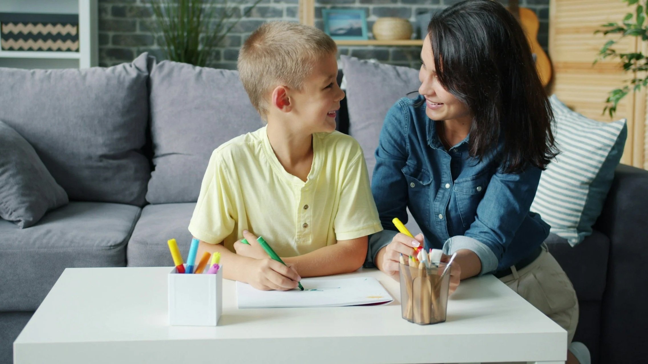 A woman and a boy smiling and looking at each other while sitting at a white table with colored markers, in a living room with a gray sofa, a brick wall, and decorative plants.