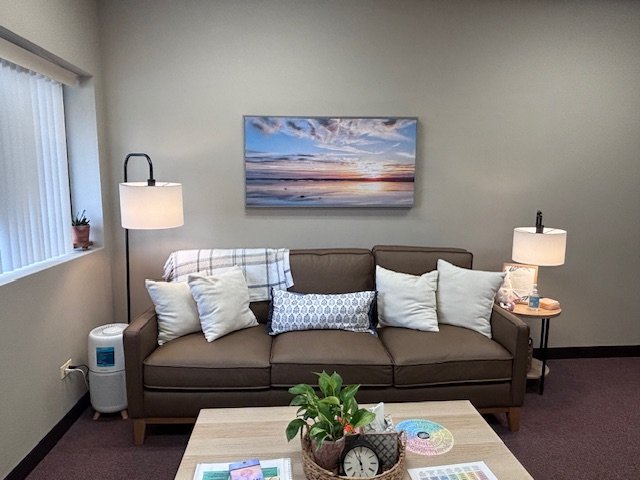 Living room with a brown sofa, white pillows, a blanket, two table lamps, a wall picture of a beach scene, a small plant on the window ledge, and a coffee table with a clock and papers.