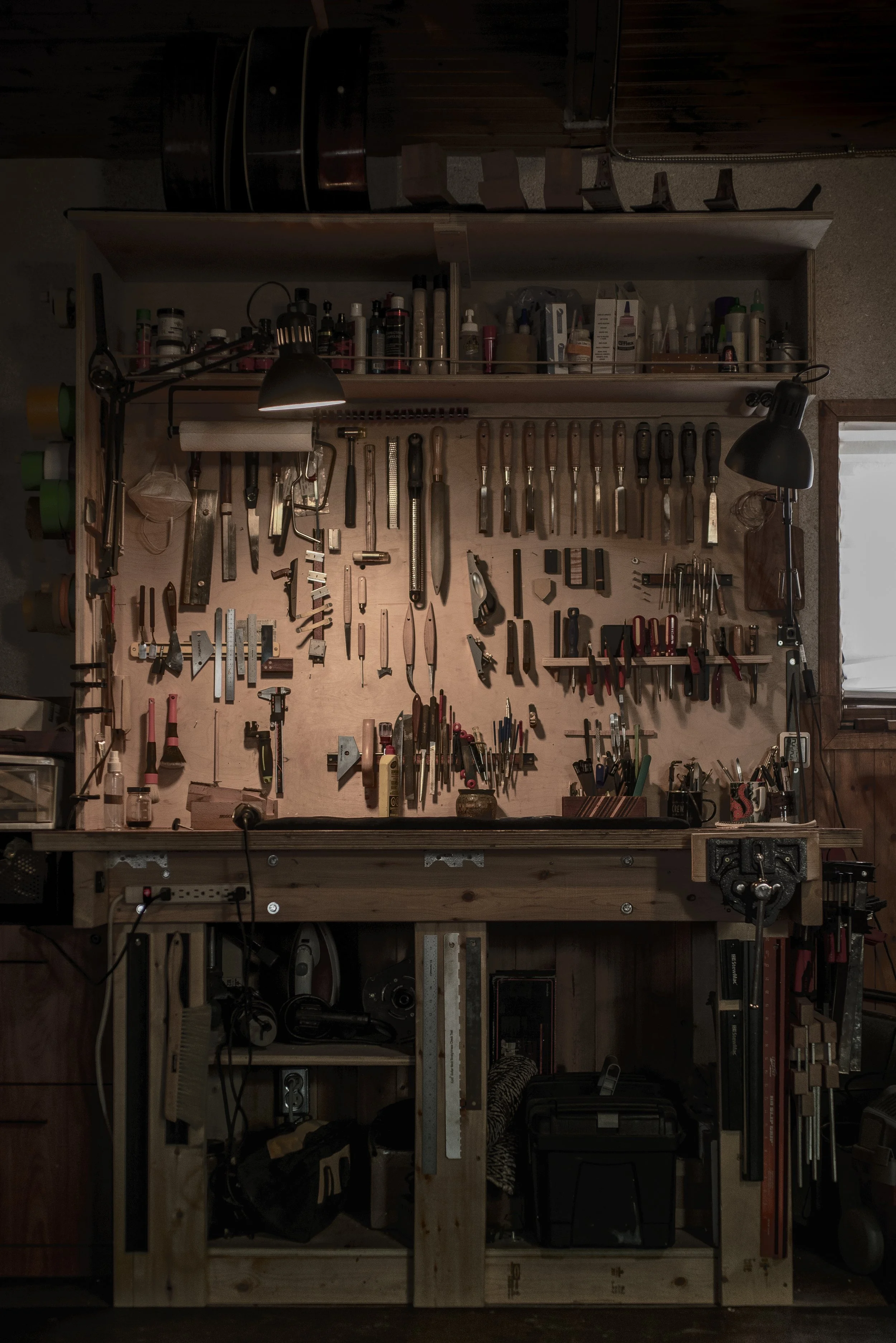 A woodworking workbench with various hand tools organized on a pegboard, including saws, chisels, hammers, and screwdrivers, with storage shelves and a window to the right.
