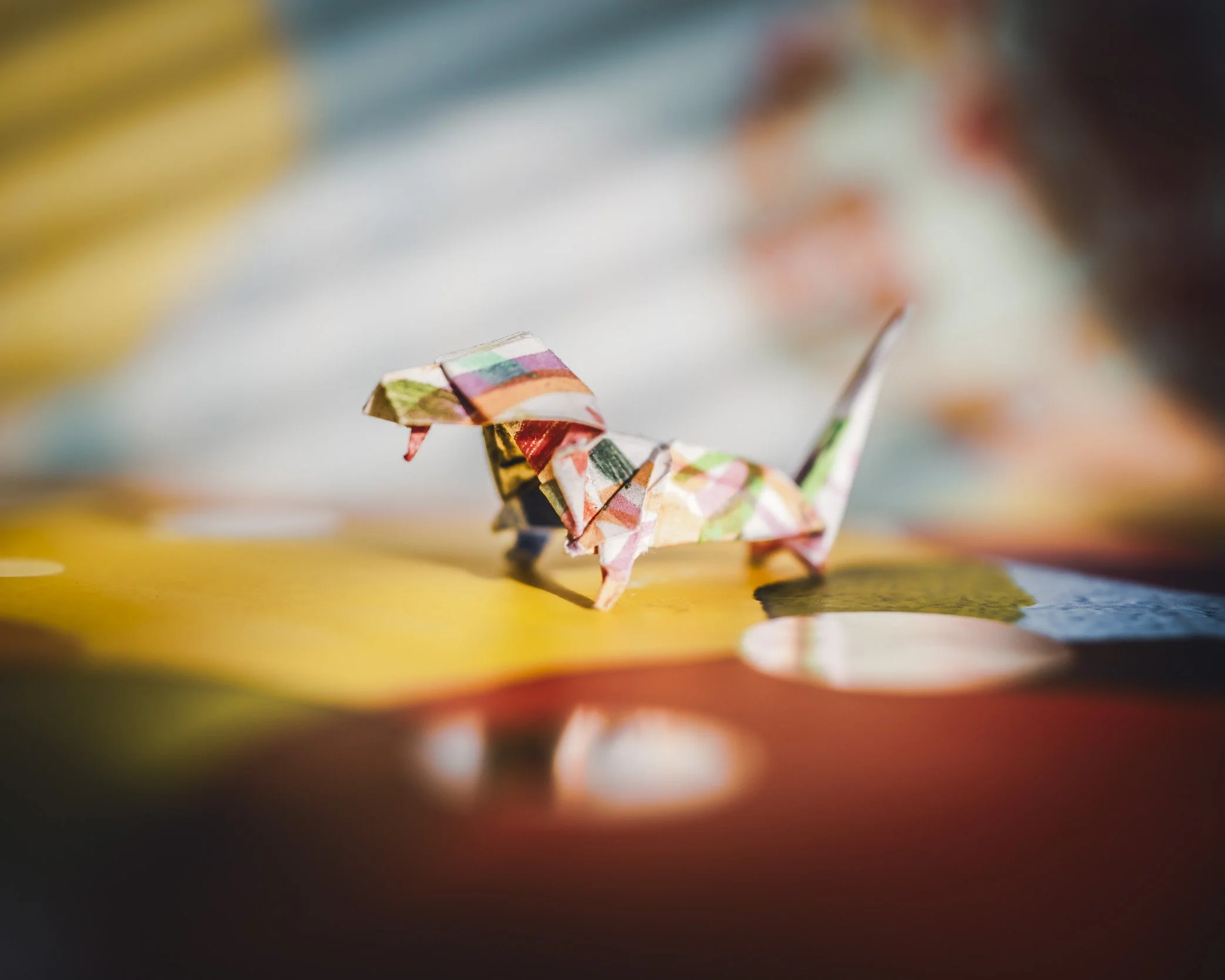 A small origami dinosaur made from colorful patterned paper on a yellow surface with a blurred background.