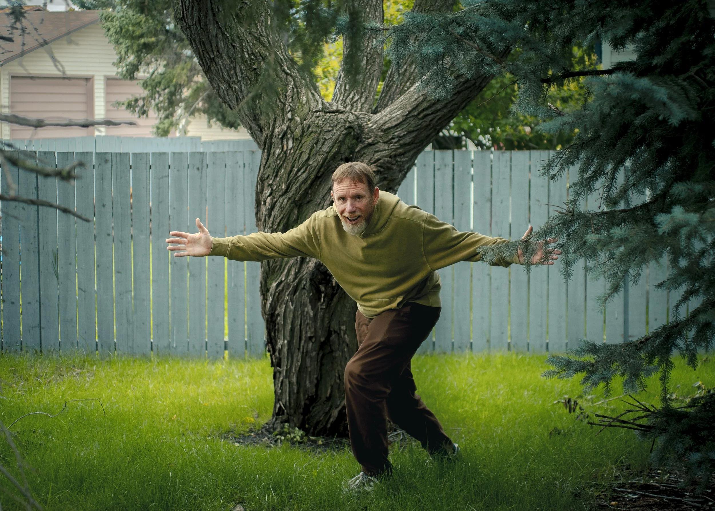 A man with short hair and a beard, wearing an olive green hoodie and brown pants, is standing in a backyard with green grass. He is leaning to one side with arms outstretched, smiling, in front of a large tree with textured bark, surrounded by other trees and a light blue wooden fence.
