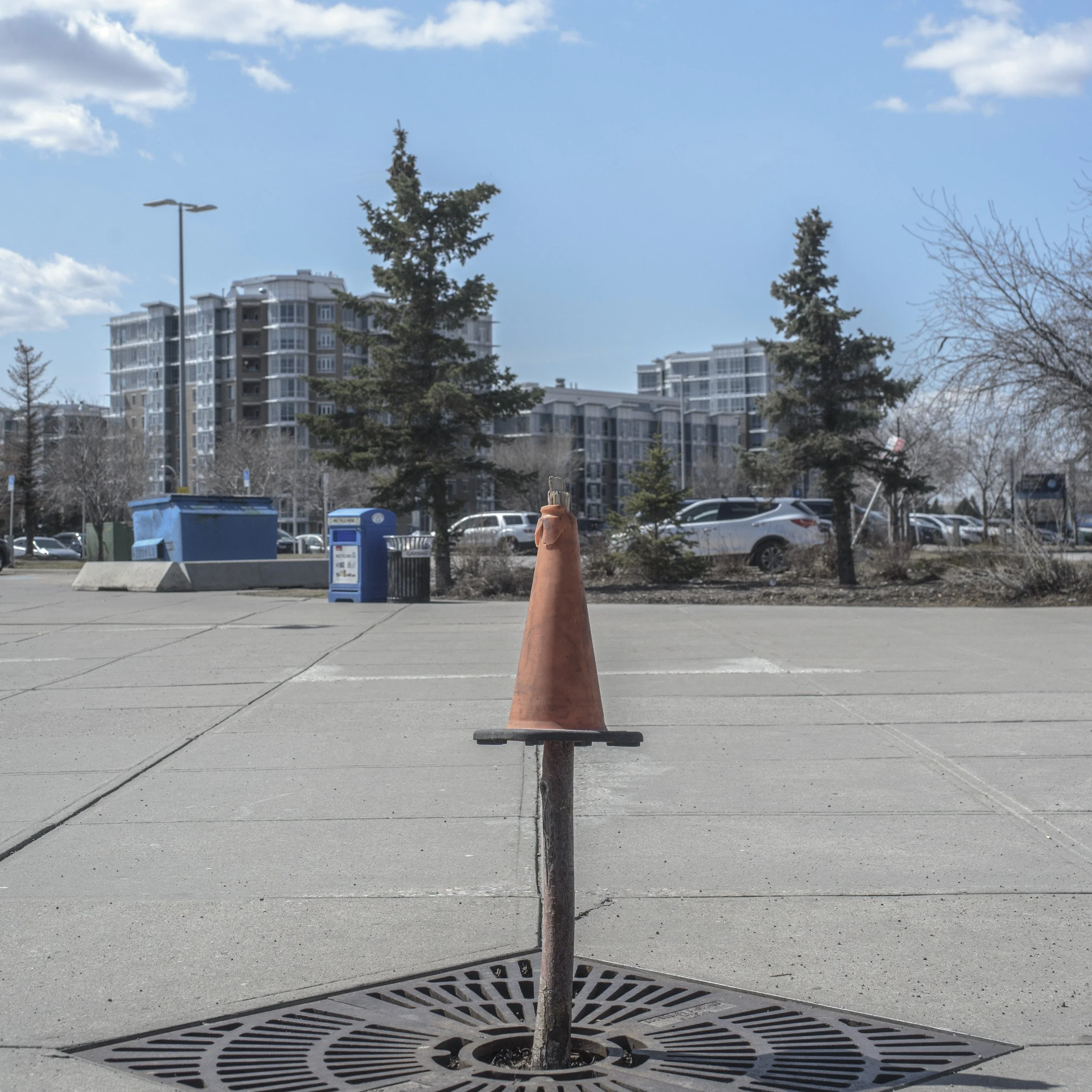 A traffic cone placed on a drain cover on a concrete parking lot with trees, parked cars, and apartment buildings in the background under a partly cloudy sky.