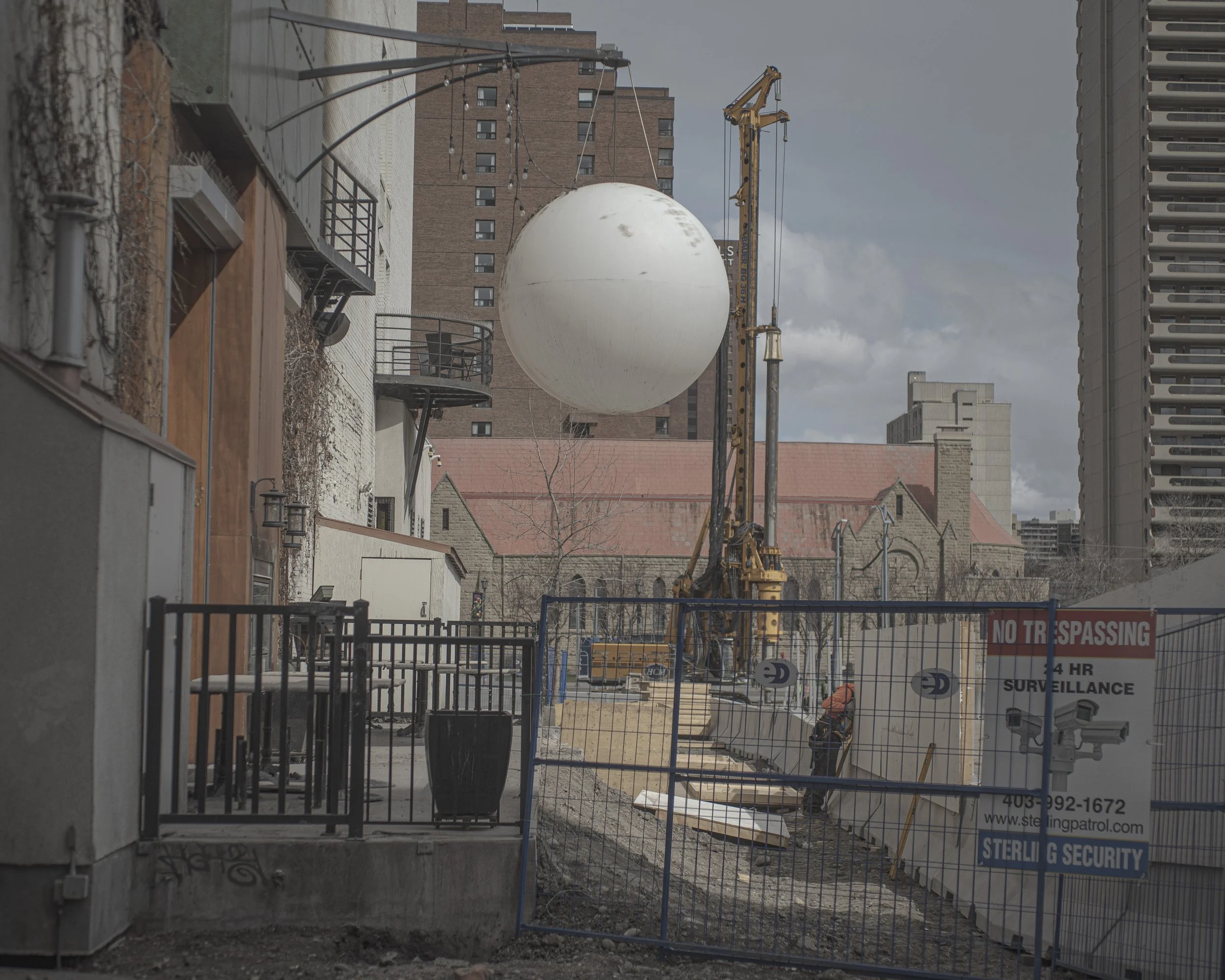 Construction site with a large white sphere, construction crane, and worker in an urban area surrounded by fencing and buildings.