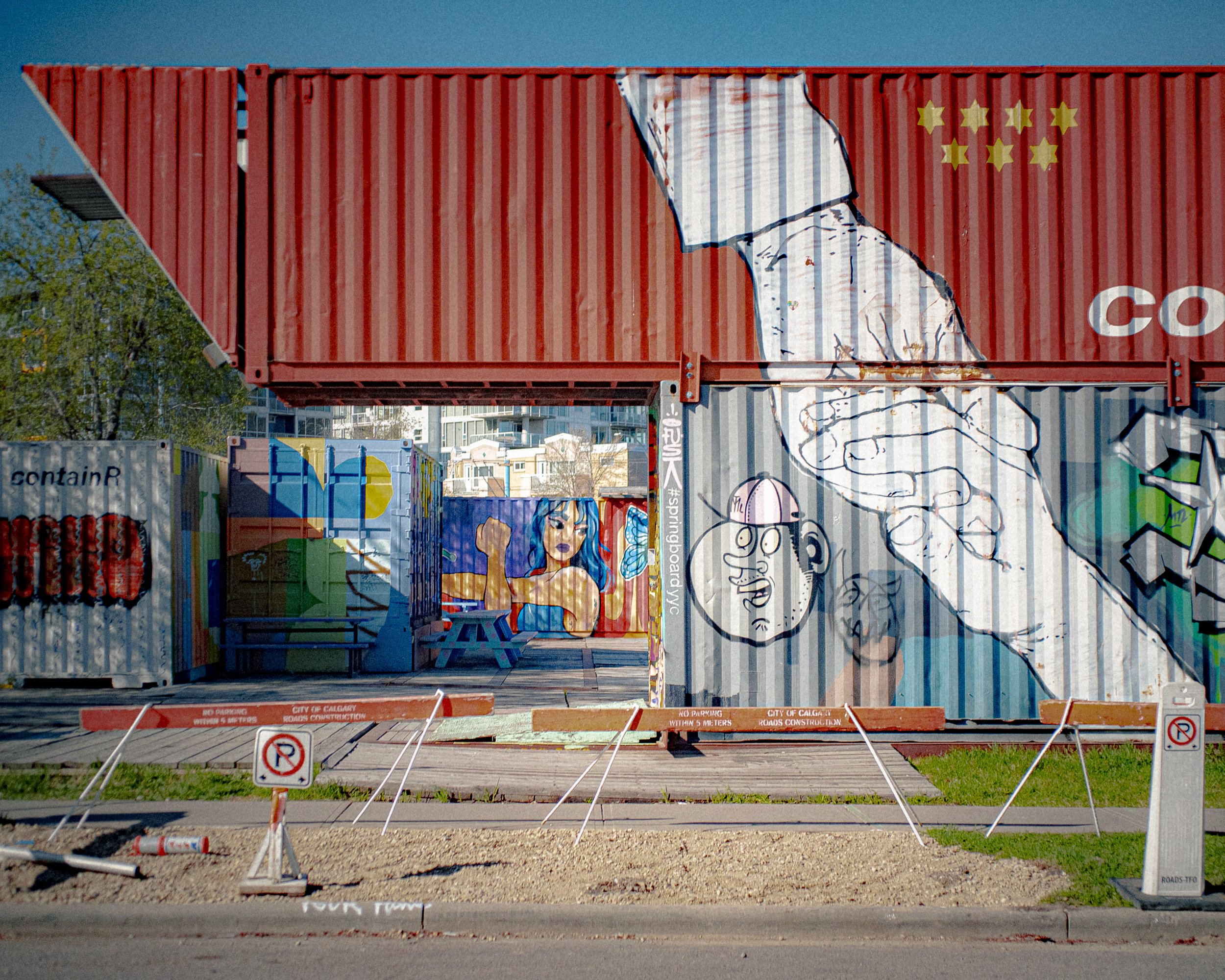 Graffiti art on shipping containers featuring a large outline of a hand holding a small figure, a portrait of a woman with blue hair, and other colorful designs, with barriers and no parking signs in front.
