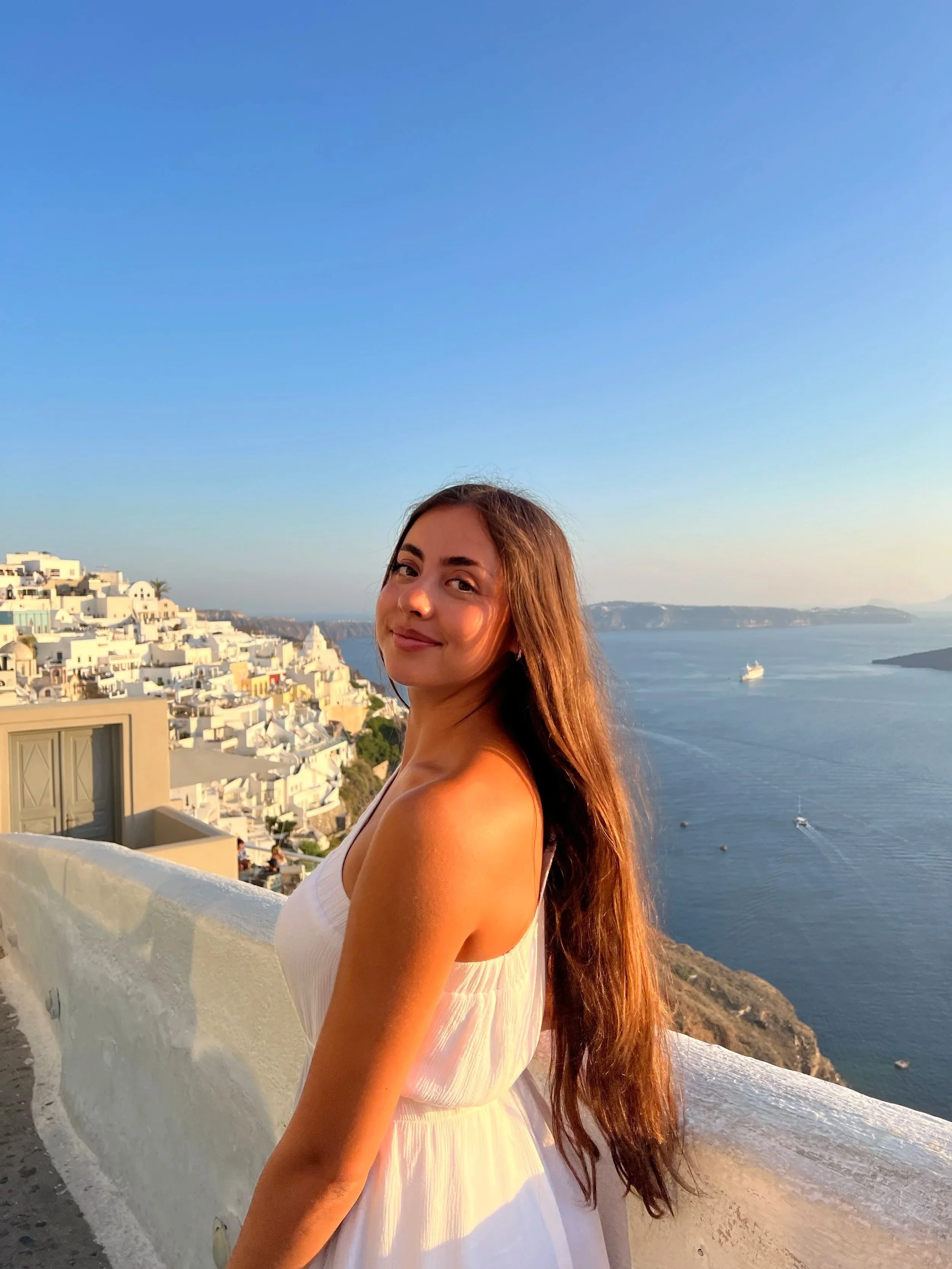A woman with long brown hair smiling at the camera, wearing a white dress, with a Mediterranean coastal town and boats on the water in the background during sunset.
