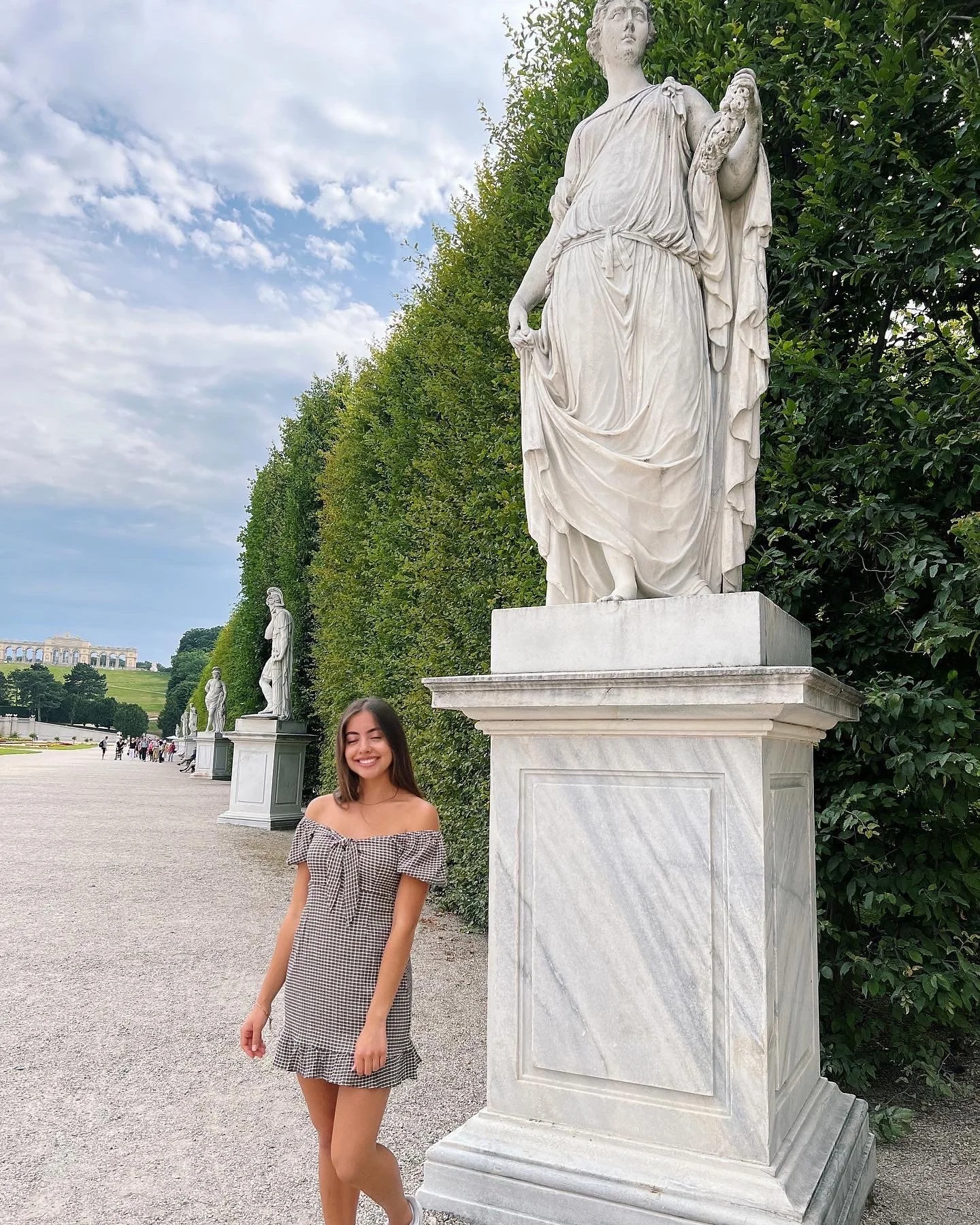 A young woman with long brown hair smiling and wearing an off-the-shoulder checkered dress standing next to a large marble statue outdoors with a row of similar statues and a green hedge in the background.