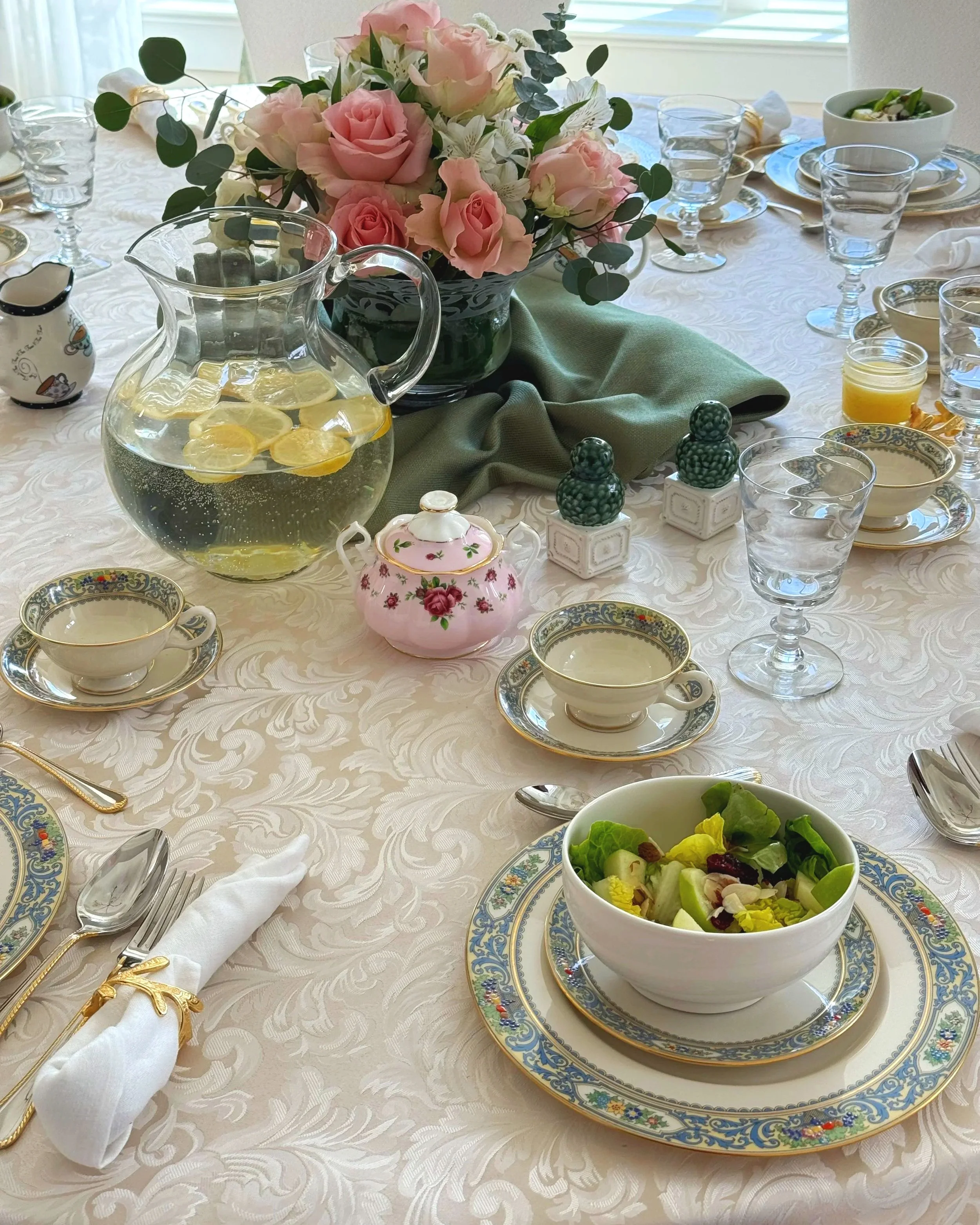 A formal dining table setup with floral centerpiece, teacups, saucers, glassware, a pitcher of lemon water, a bowl of salad, salt and pepper shakers, a small pink teapot, and a china plate with a napkin and utensils.