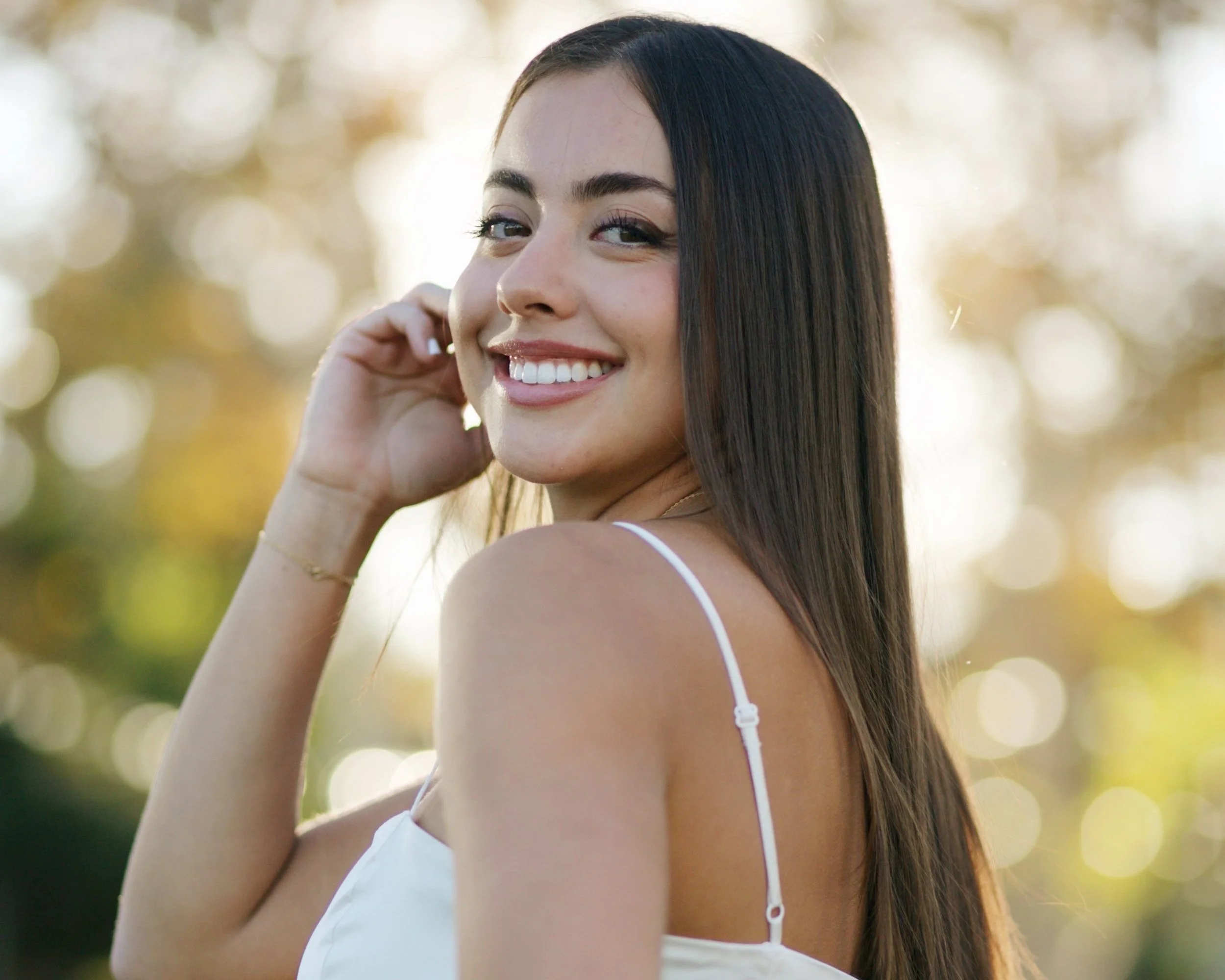 Jessi, a smiling woman with long hair wearing a silky white dress, outdoors with blurred background.