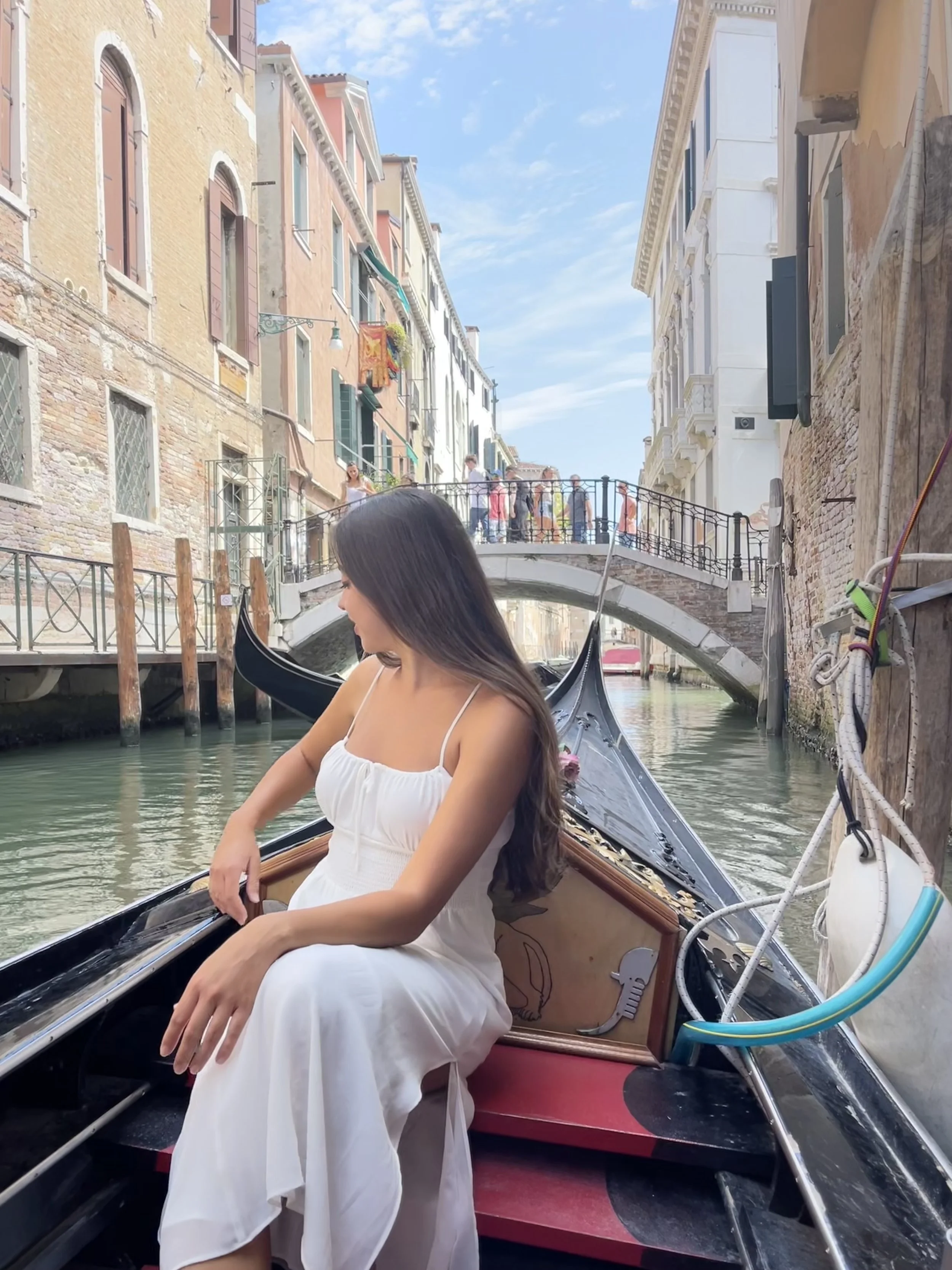 A woman in a white dress sitting in a gondola on a canal in Venice, Italy, with colorful buildings, a small bridge, and pedestrians in the background.