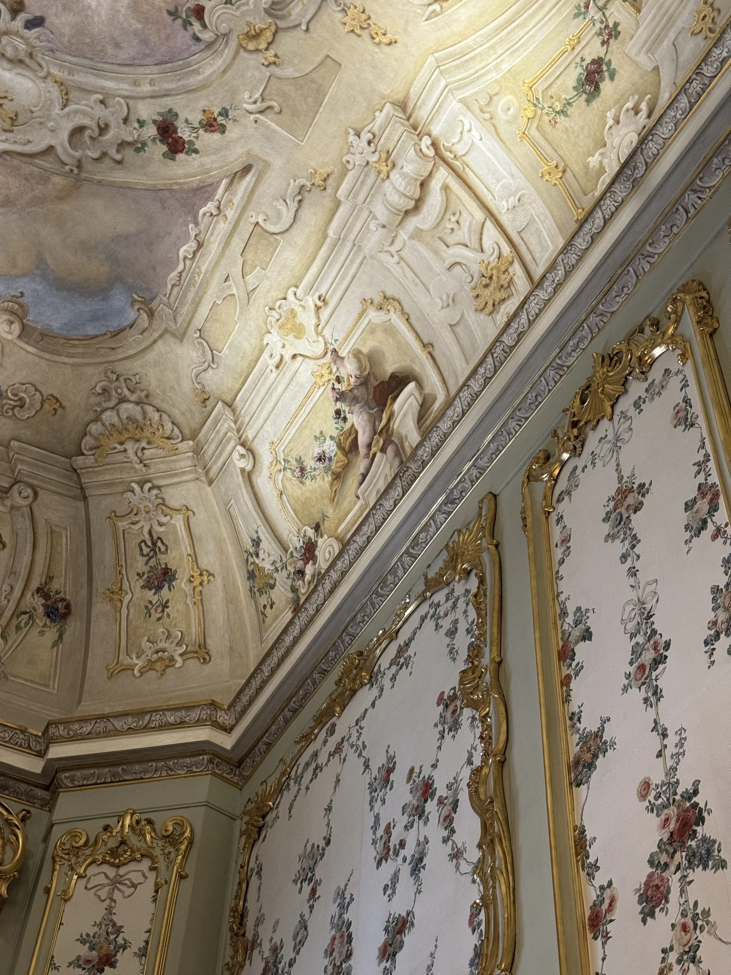 Ornate ceiling with elaborate gold and white molding, floral motifs, and a cherub figure surrounded by flowers.