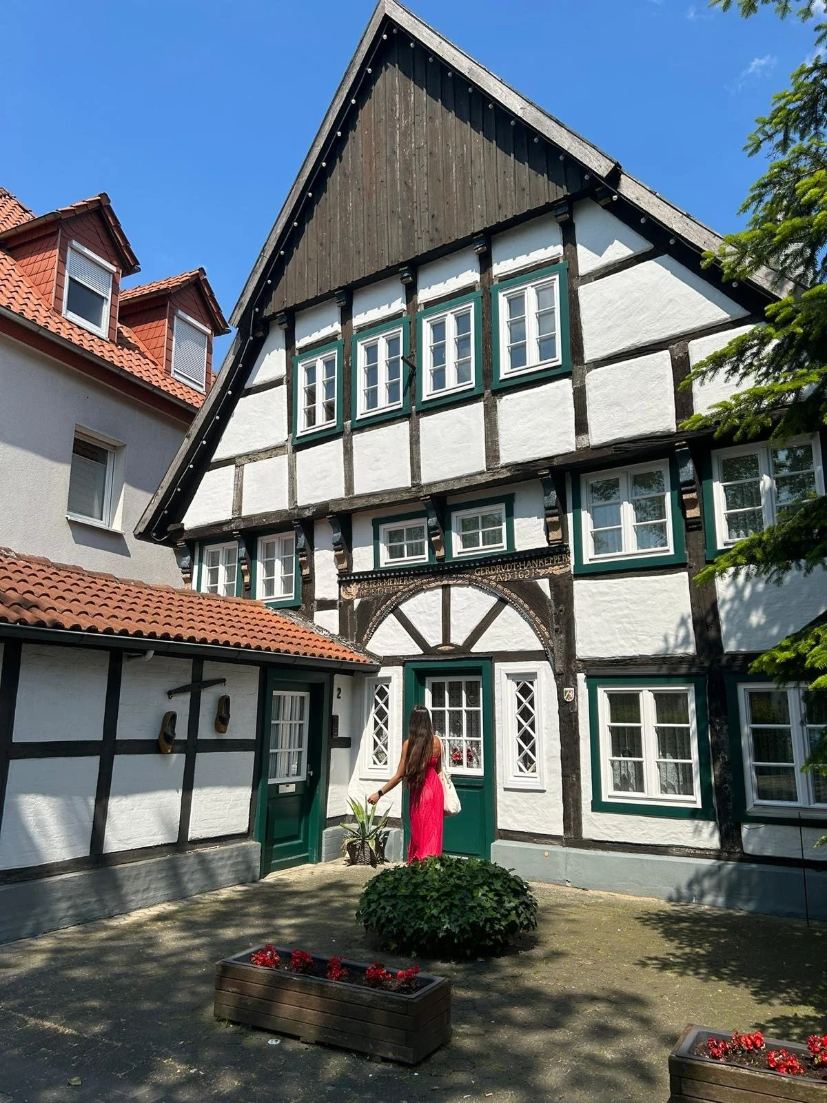 A woman in a pink dress standing in front of a traditional German-style house with a painted timber frame, white walls, green window frames, and a red-tiled roof, with garden plants and flower boxes.