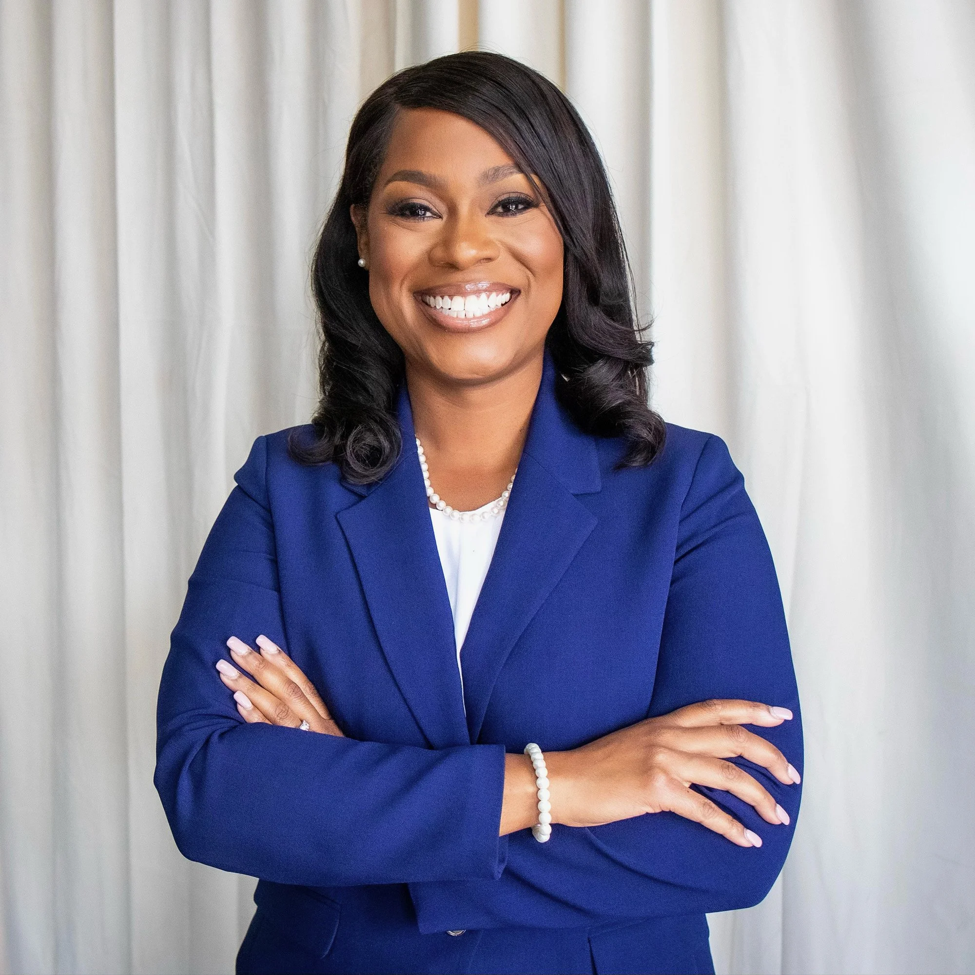 A professional woman in a blue blazer and white top, smiling and crossing her arms, standing in front of white curtains.