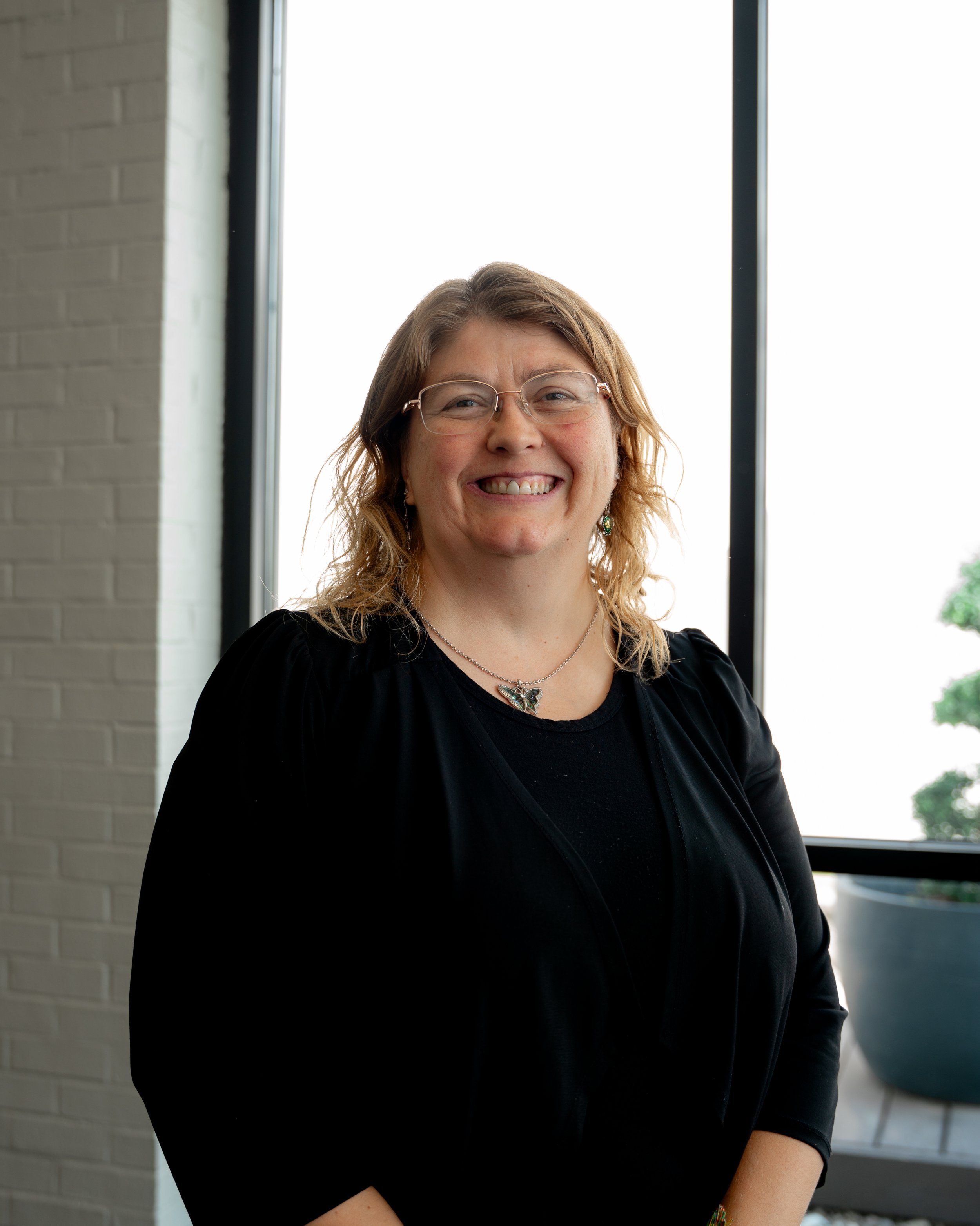Portrait of a smiling woman with glasses standing indoors near a window, wearing a black top and jewelry.