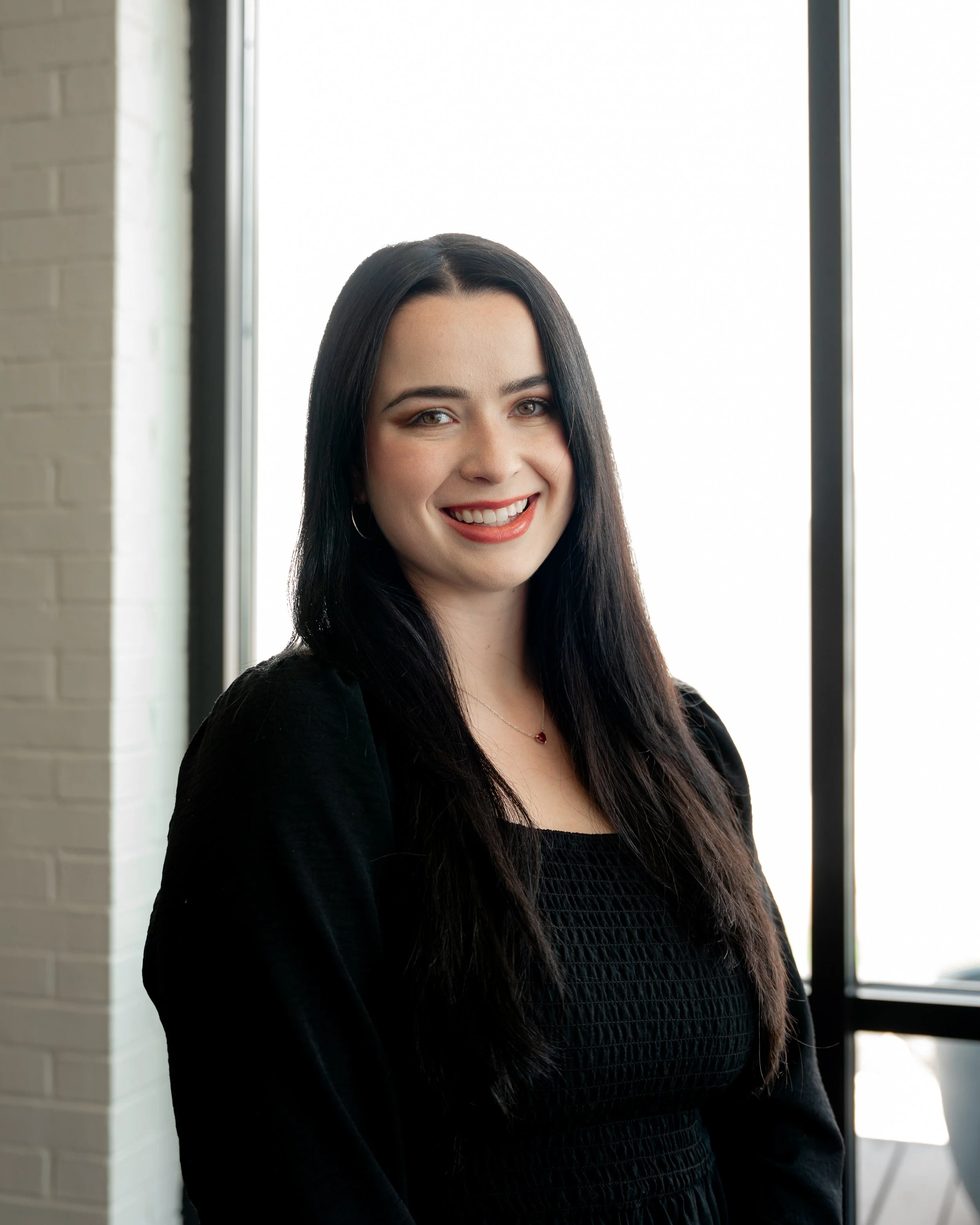 A woman with long dark hair smiling at the camera, standing inside near a window with a brick wall visible on her left.