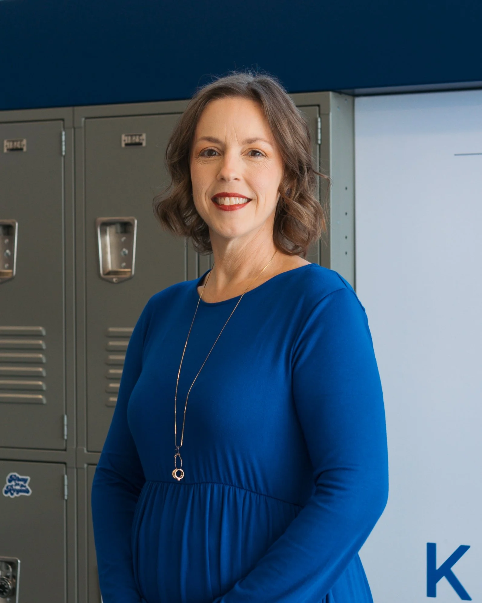 A woman with shoulder-length brown hair and red lipstick, wearing a royal blue long-sleeve dress with a necklace, standing in front of gray lockers in a school hallway.