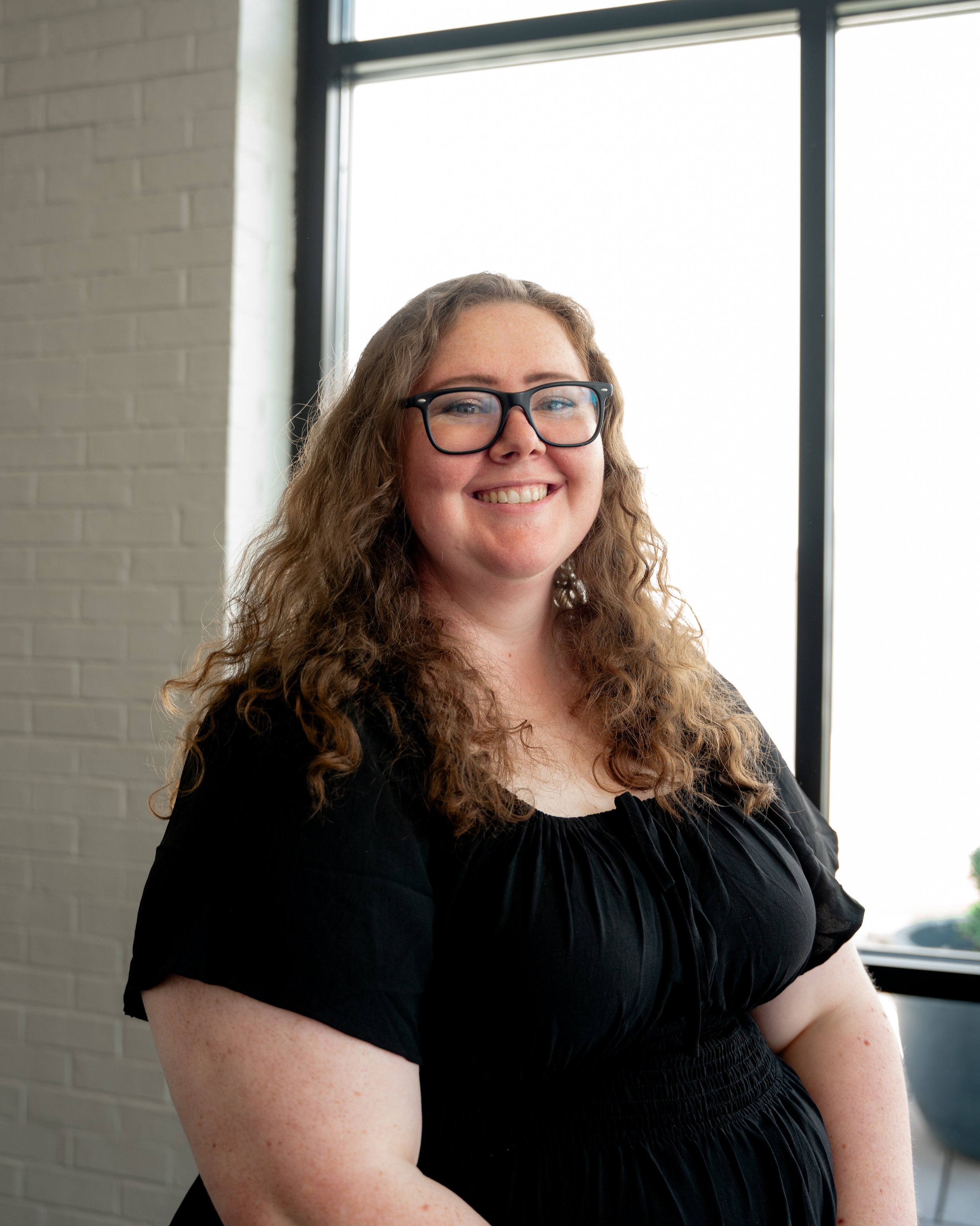 A woman with glasses and curly hair smiling in front of a large window with a brick wall in the background.