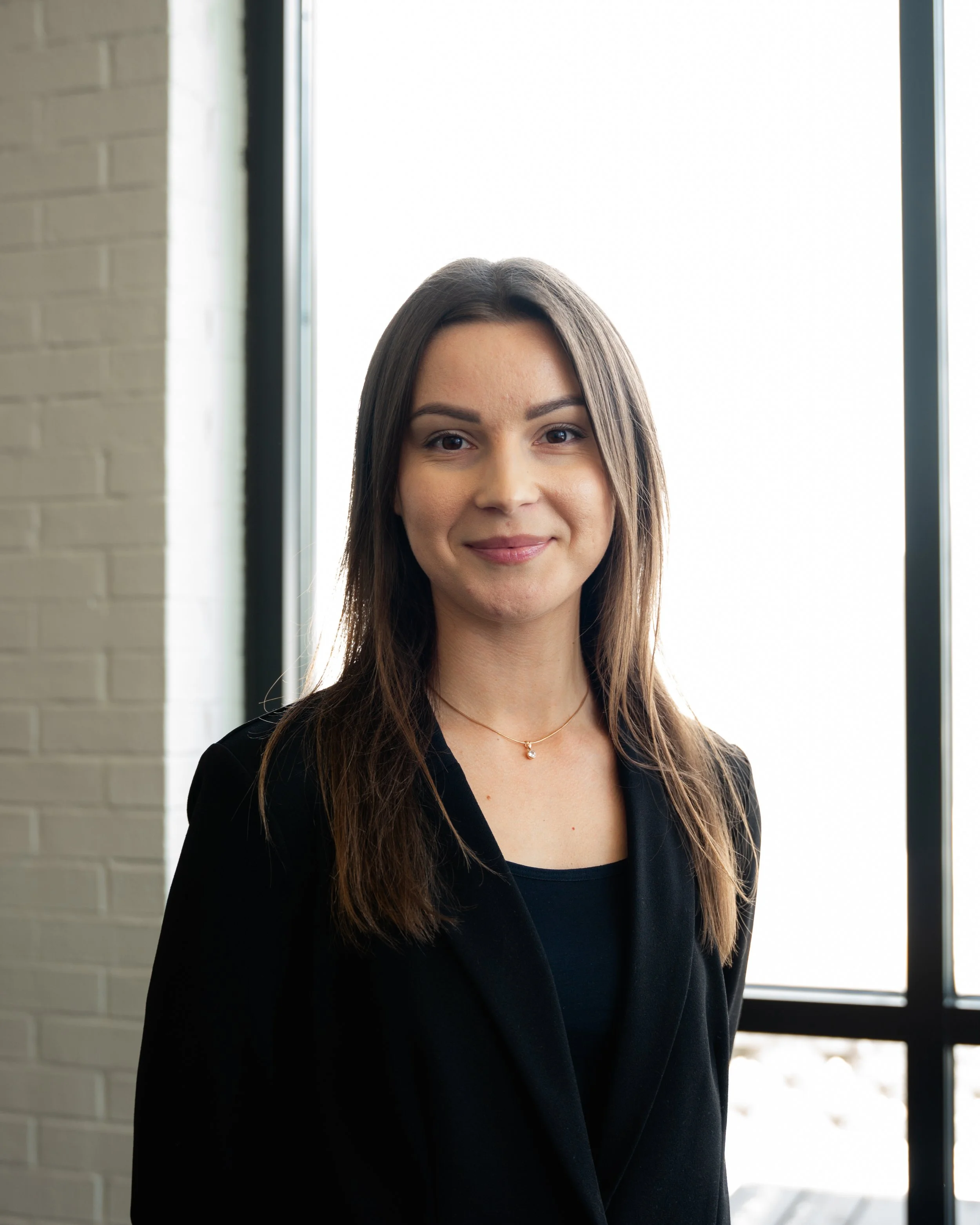 A young woman with brown hair and a black blazer standing indoors by large windows, smiling at the camera.