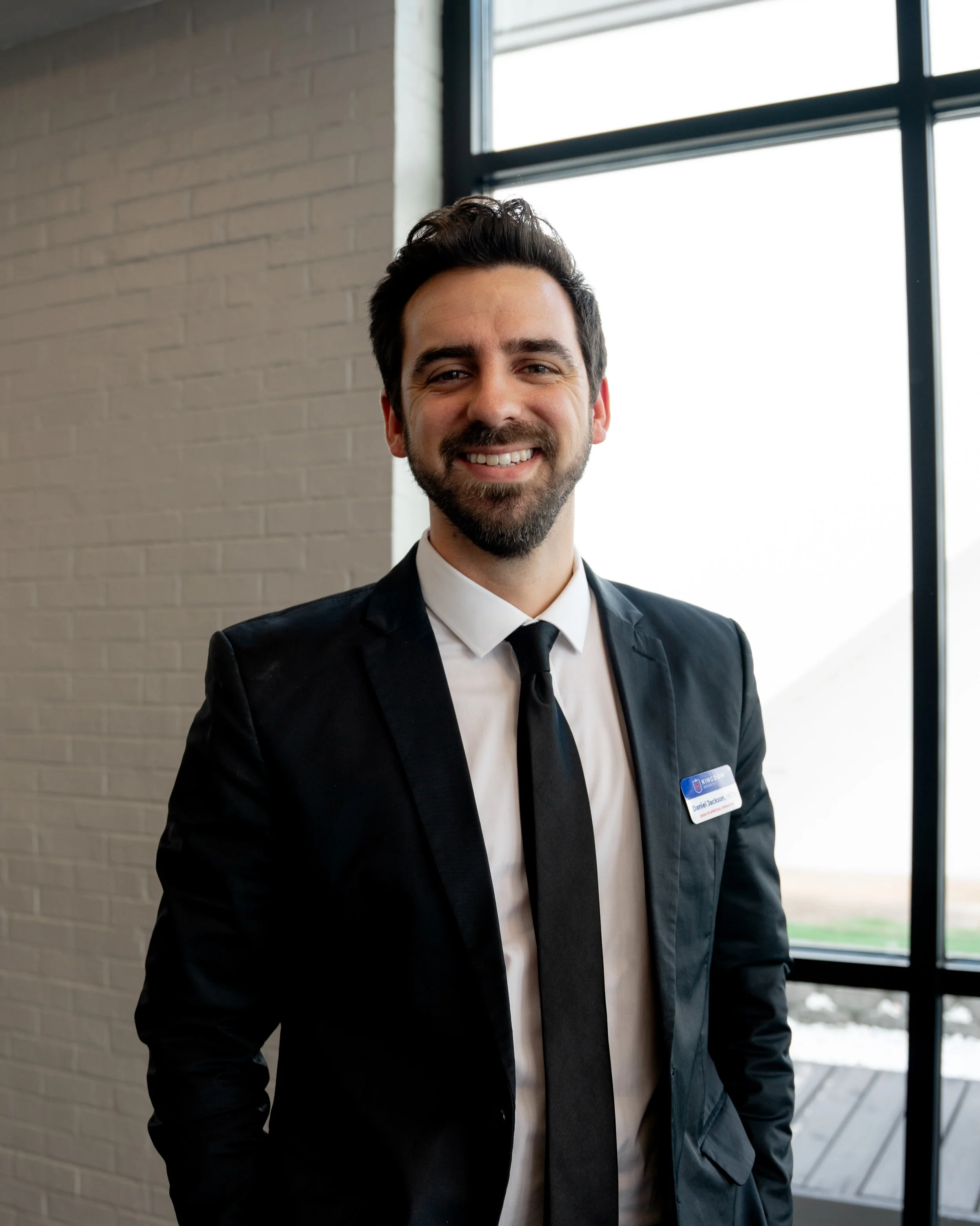 Smiling man in a black suit with a name badge, standing inside near a large window.
