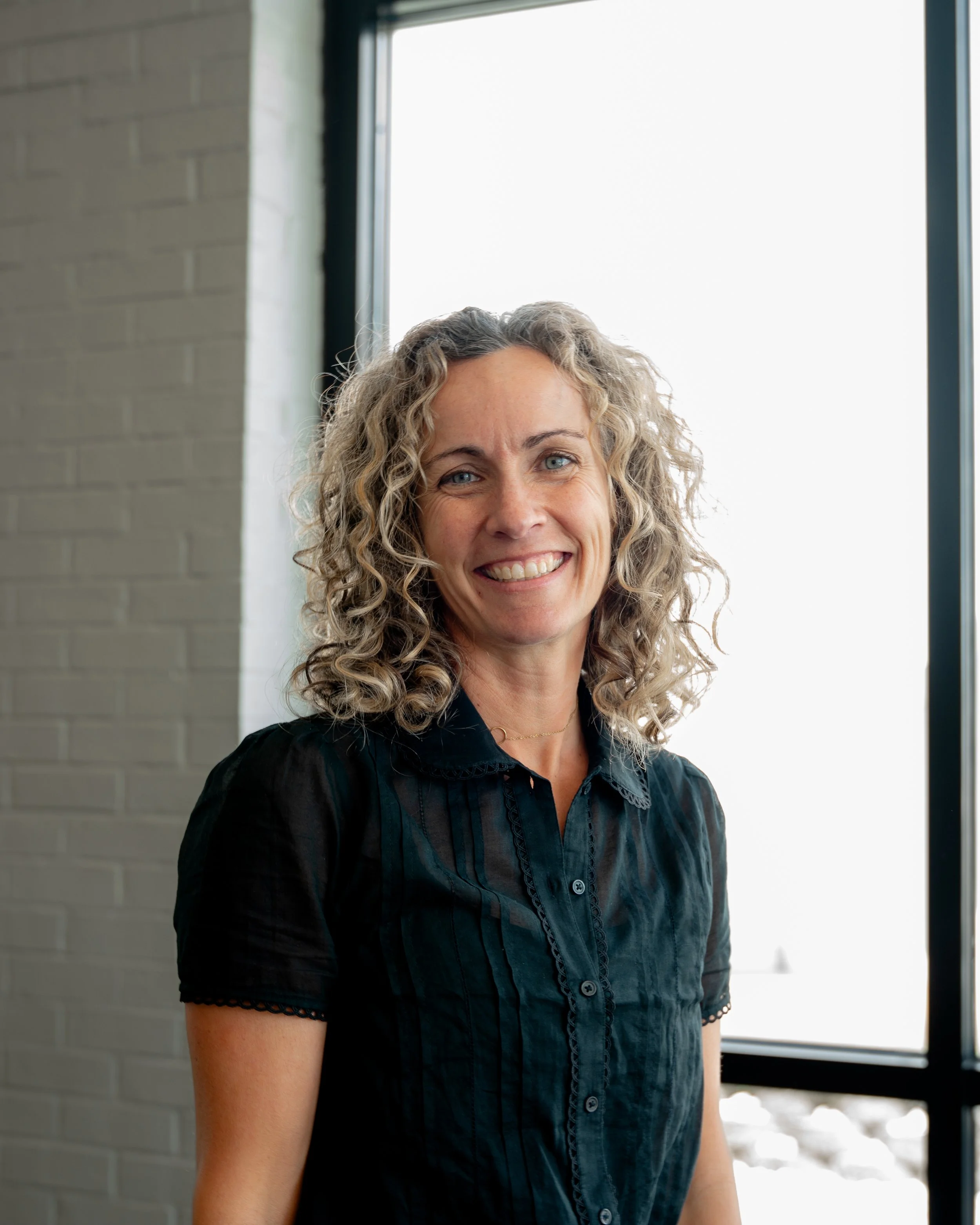 A woman with curly blonde hair and a black blouse standing indoors near a large window, smiling at the camera.