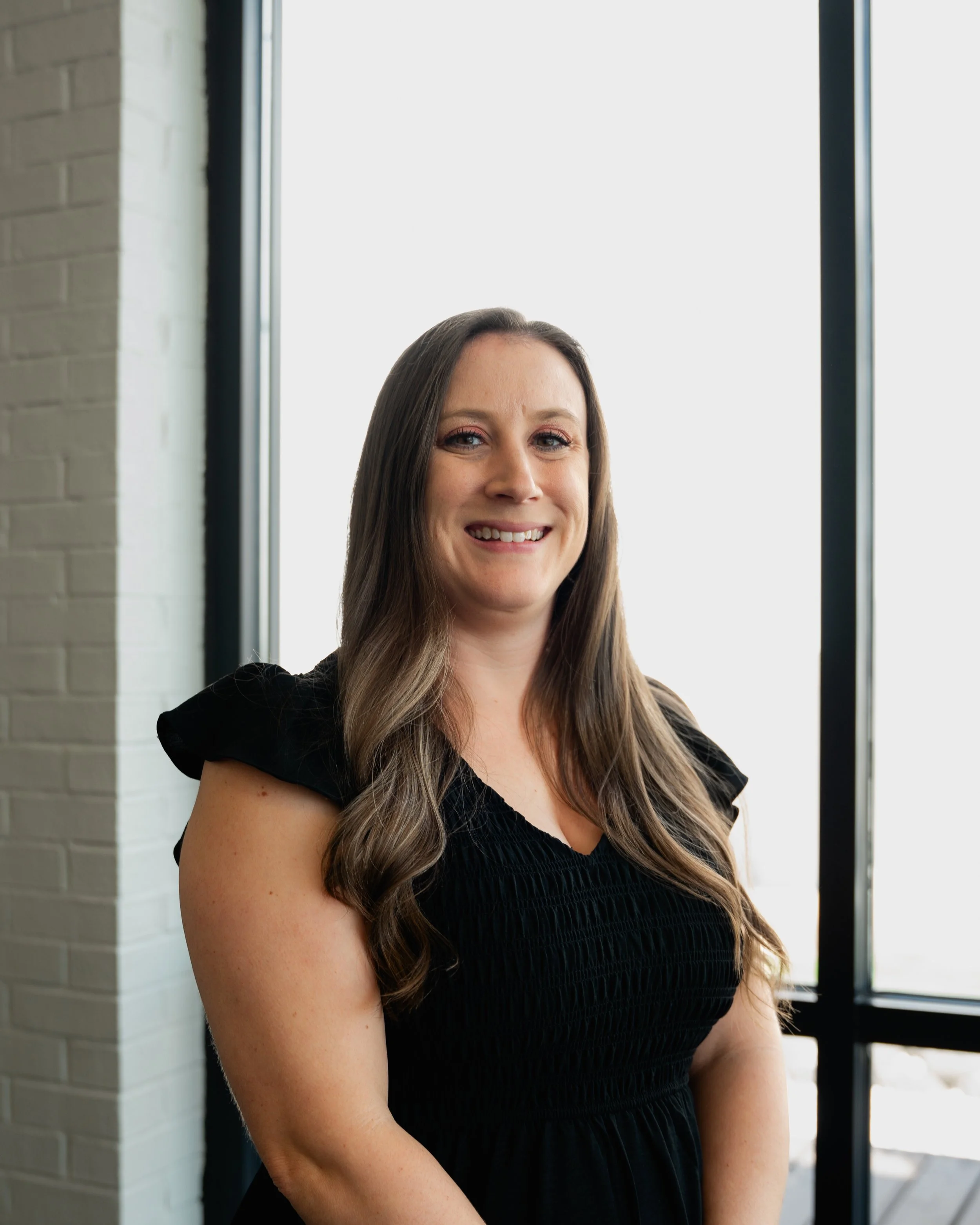 A woman with long brown hair smiling, standing near a large window with a brick wall to her left.