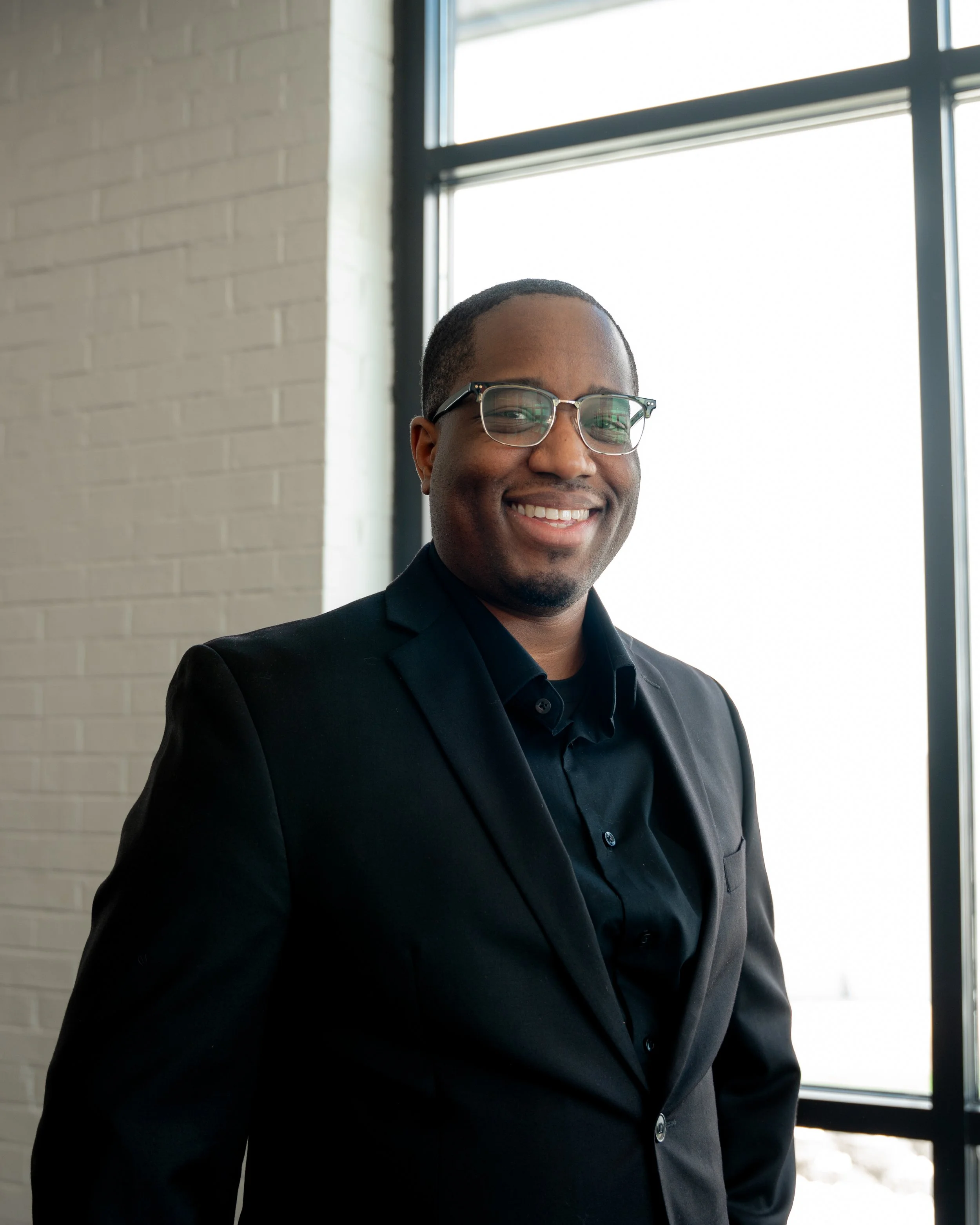 A smiling man wearing glasses and a black suit jacket standing indoors near a large window with natural light.
