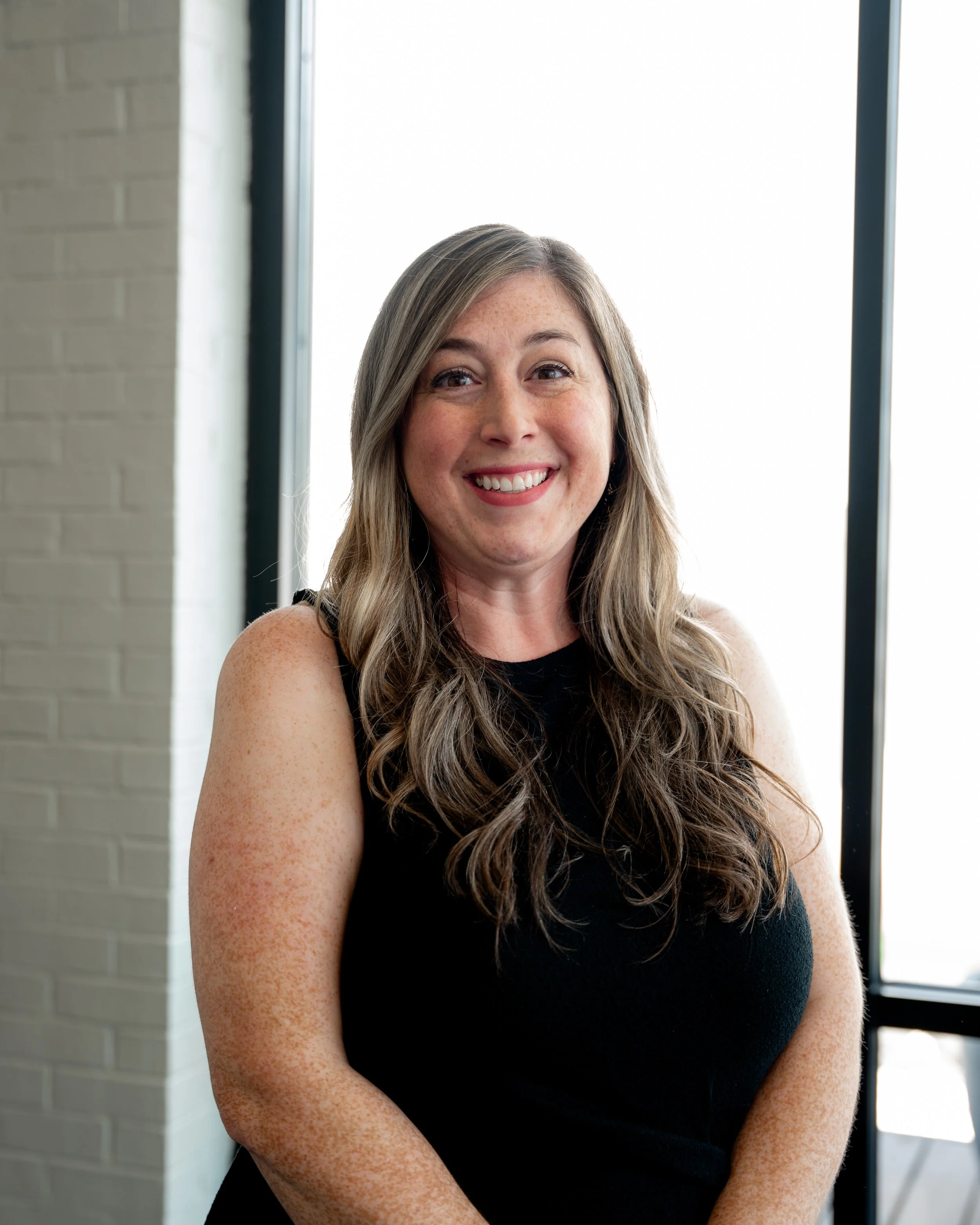 A woman with long, wavy hair and freckles, smiling and wearing a black sleeveless top, standing indoors near a large window with a white brick wall in the background.