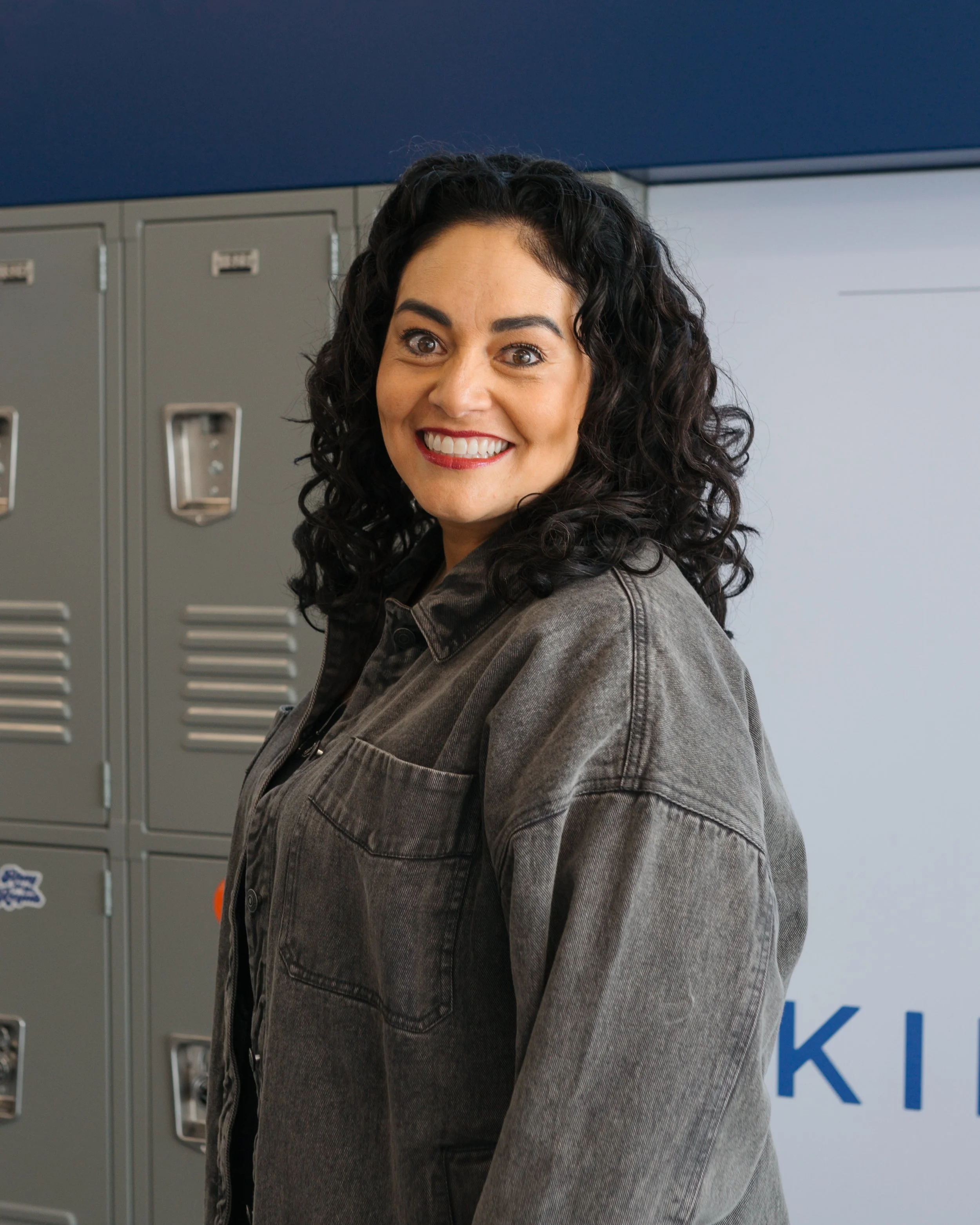 A woman with curly dark hair smiling in front of lockers.