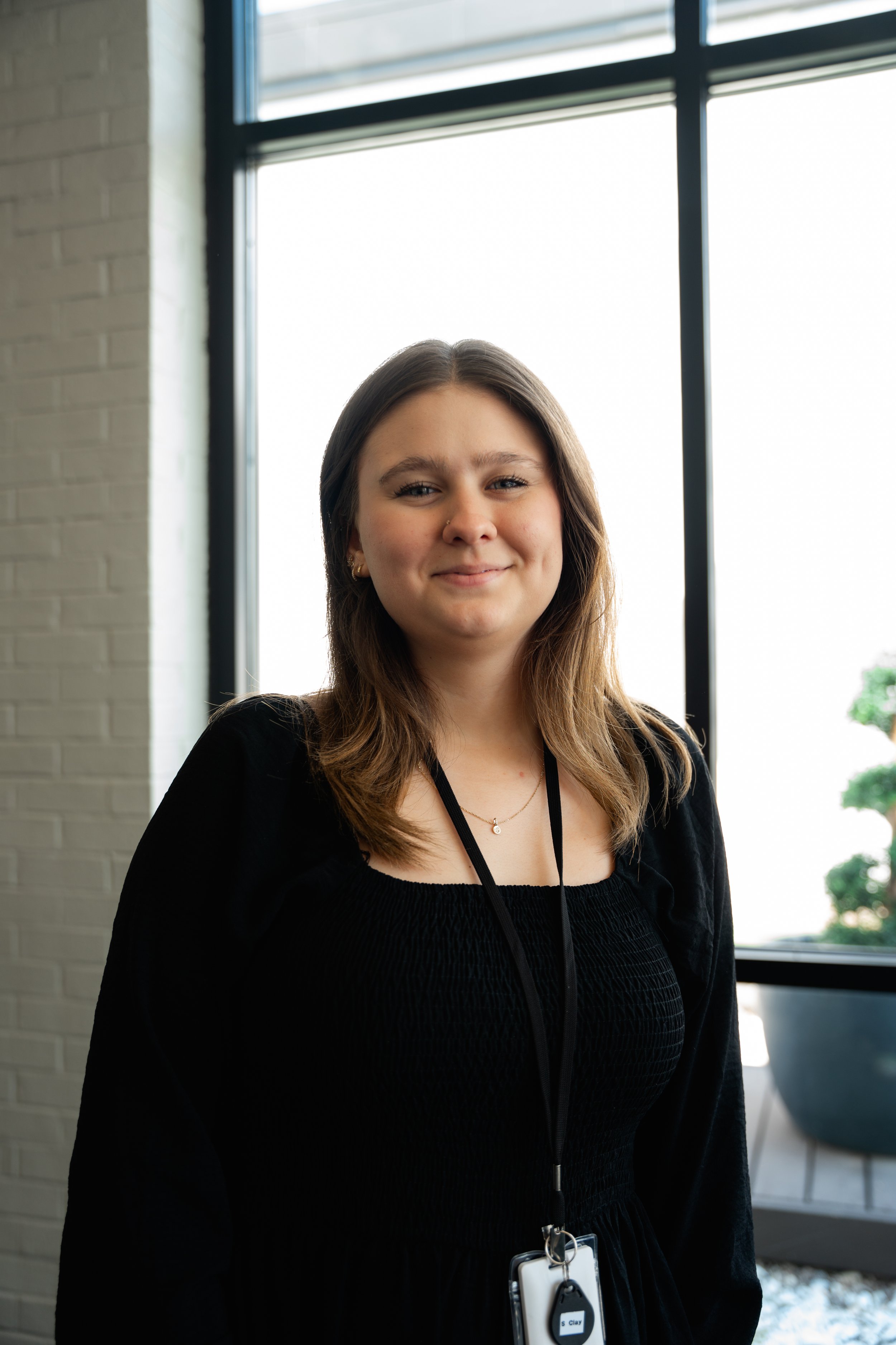 A young woman with shoulder-length brown hair, smiling, standing indoors in front of a large window with black framing, wearing a black dress and a lanyard with an ID badge.