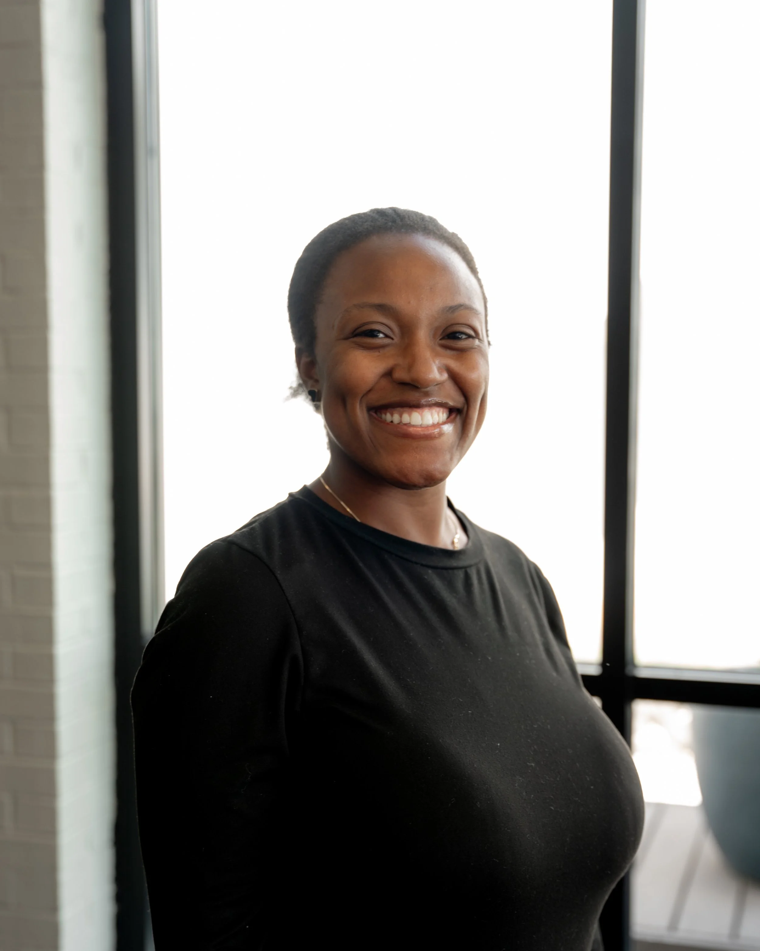 A smiling woman with dark hair pulled back, wearing a black shirt, standing by a large window.