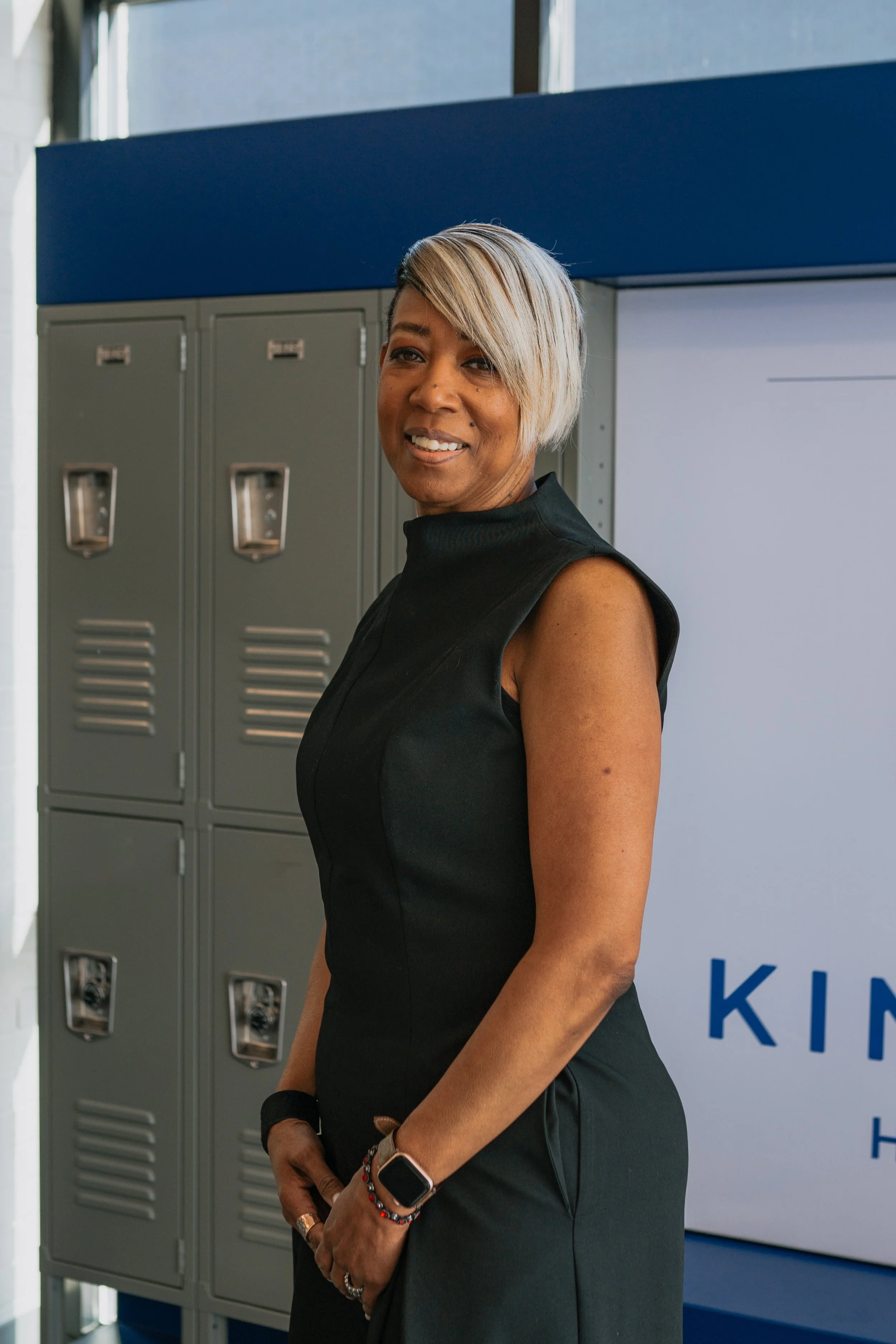 Woman with short, blonde and black hair, wearing a sleeveless black dress and smartwatch, standing in front of grey lockers and a white wall with part of a sign visible.