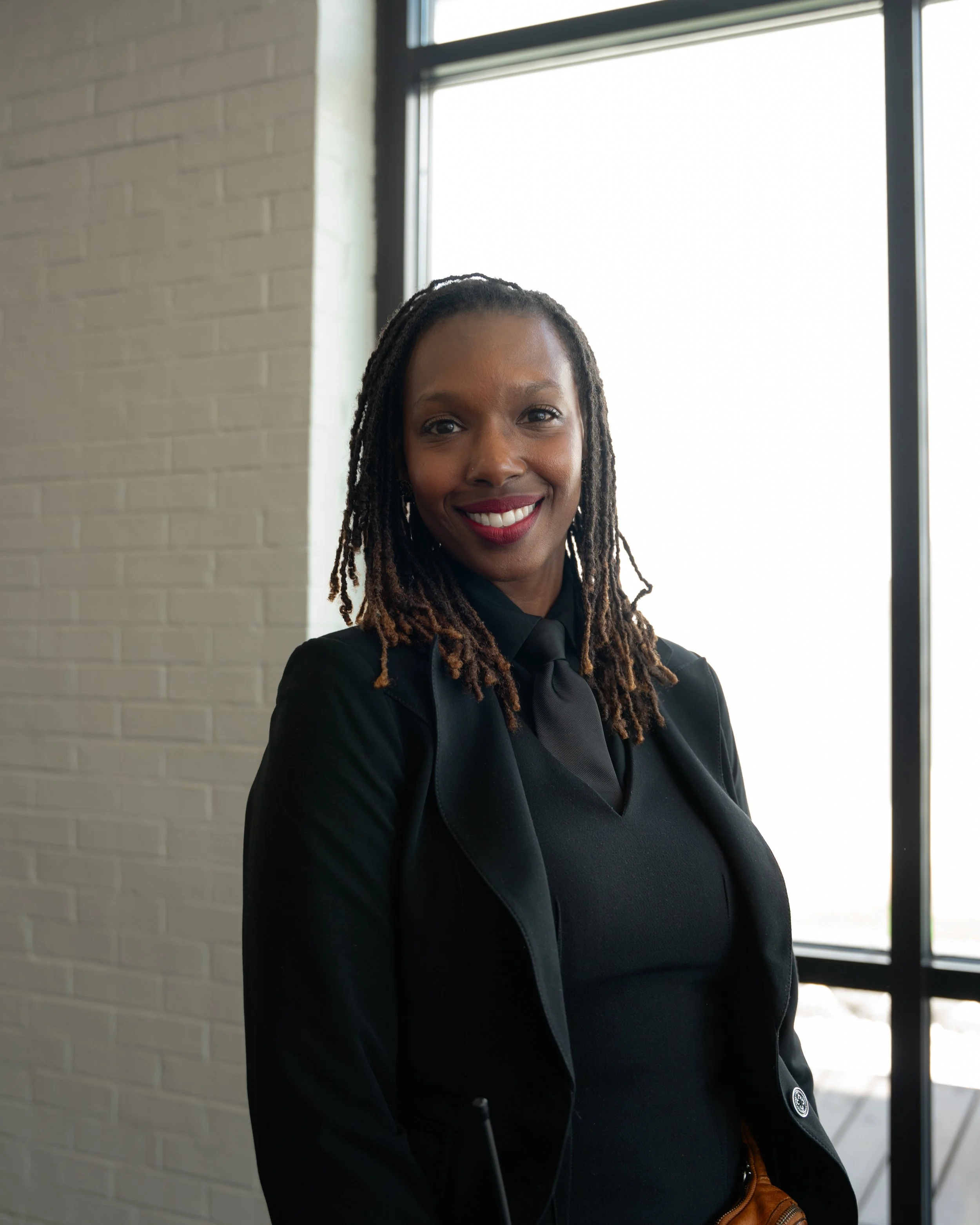 A woman with braids, wearing a black blazer and black top, smiling indoors near a large window with a brick wall behind her.