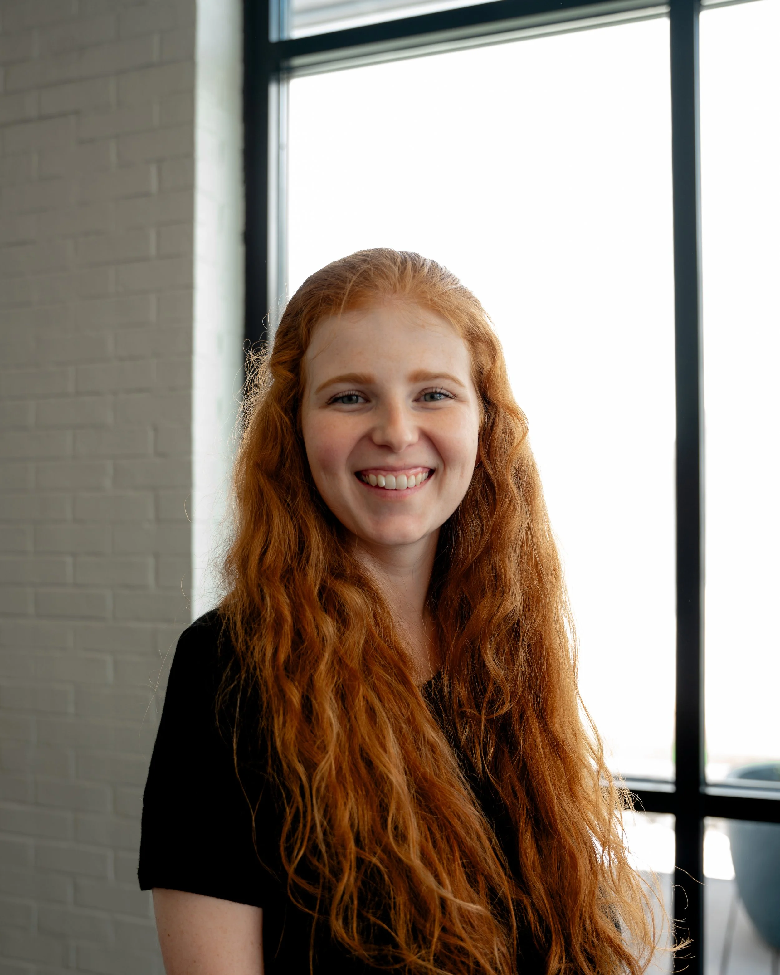 A young woman with long, curly red hair and fair skin smiling indoors near a window with a white brick wall in the background.