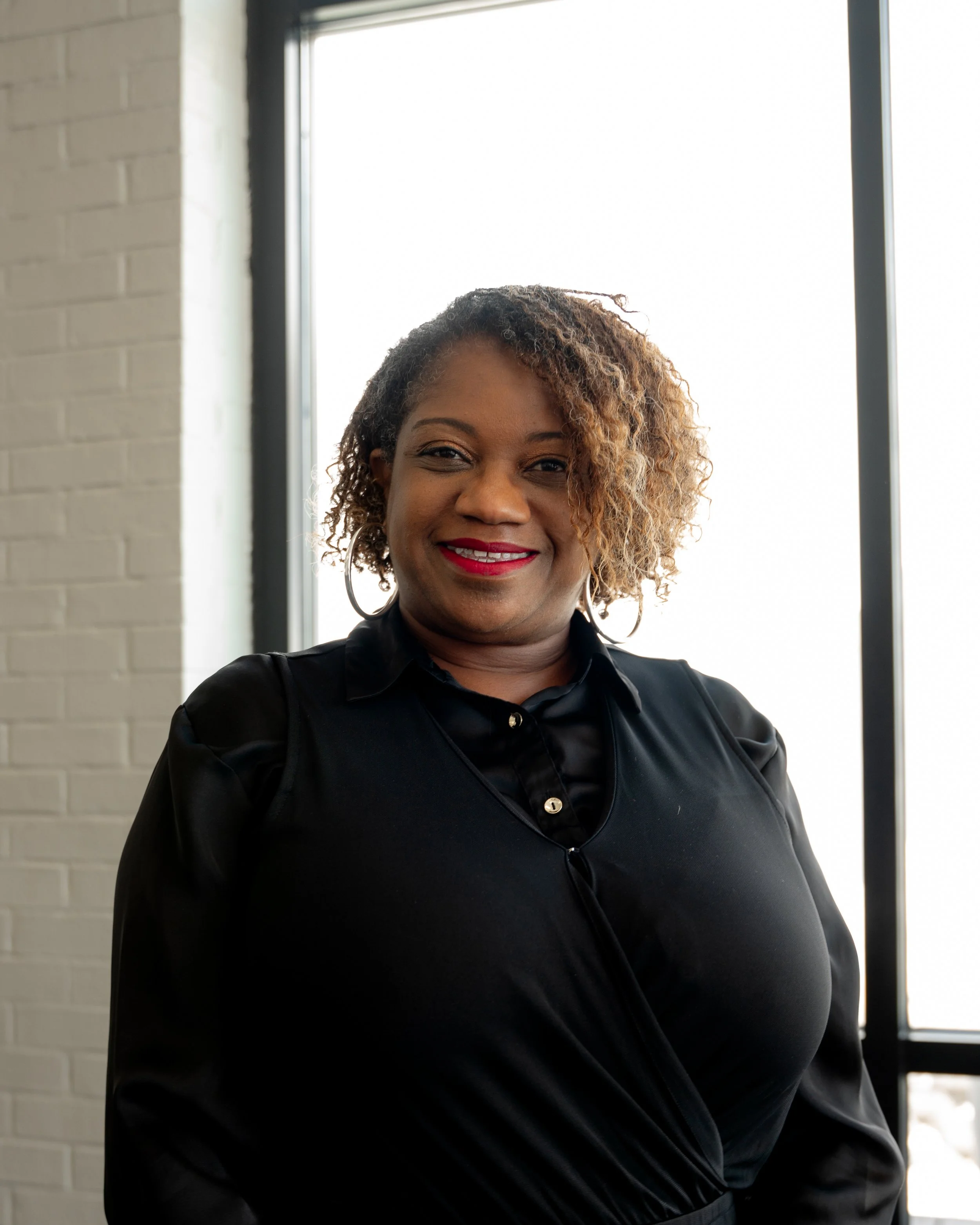 A smiling woman with light brown, curly hair, wearing a black blouse, standing in front of a large window with bright light. Brick wall is visible on the left side.