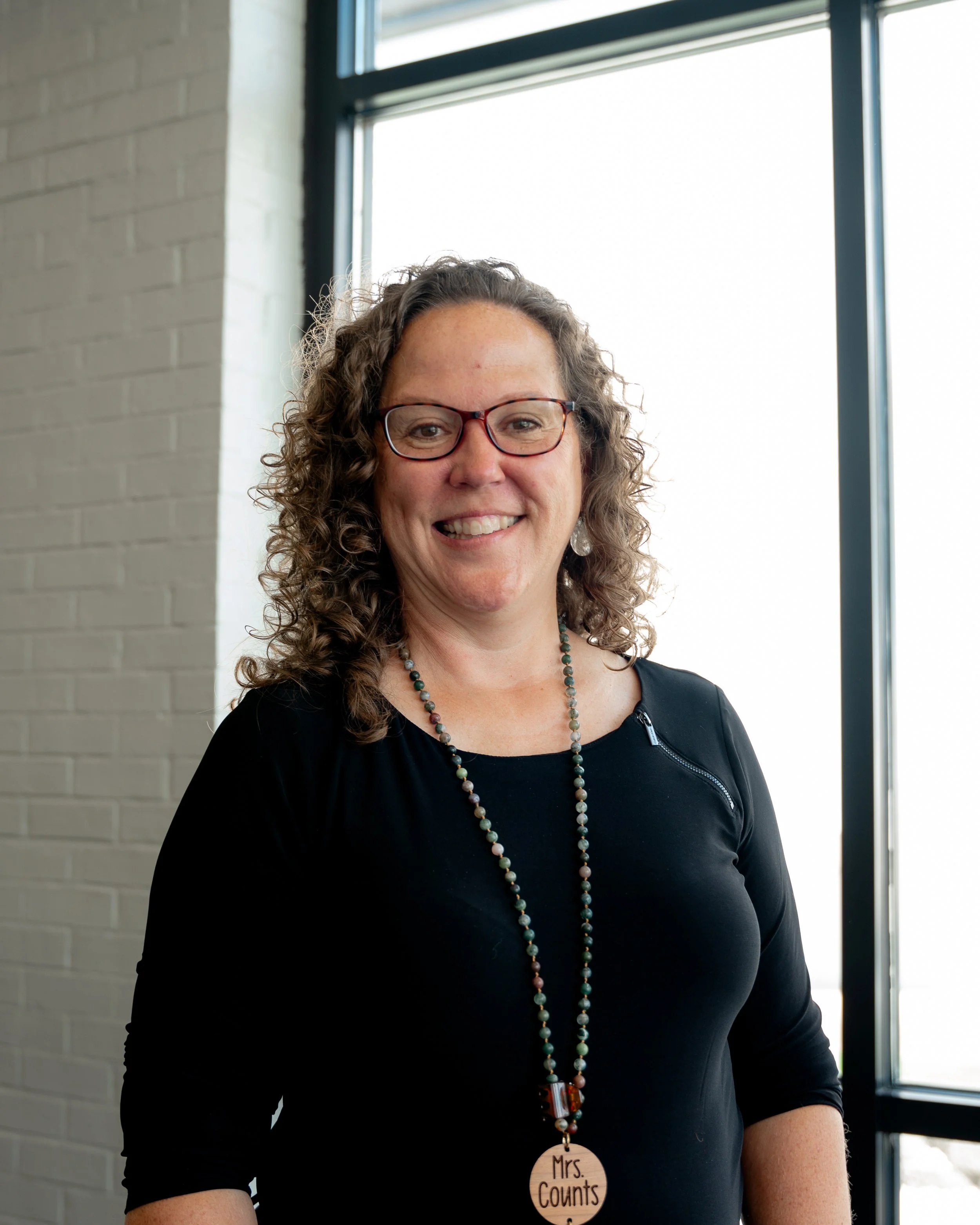 A woman with curly brown hair, wearing glasses, a black top with a zipper on the shoulder, and a beaded necklace with a wooden pendant that says 'Mrs. Counts'.