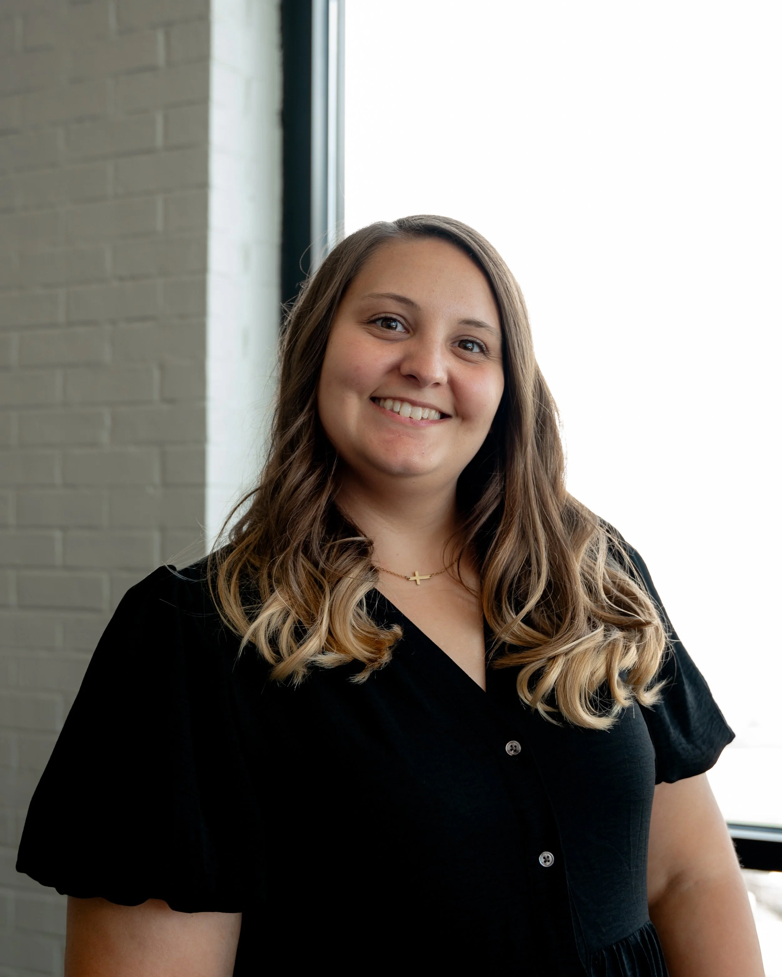 A young woman with long, wavy, light brown hair, smiling, wearing a black blouse and a small gold cross necklace, standing indoors near a window with natural light.