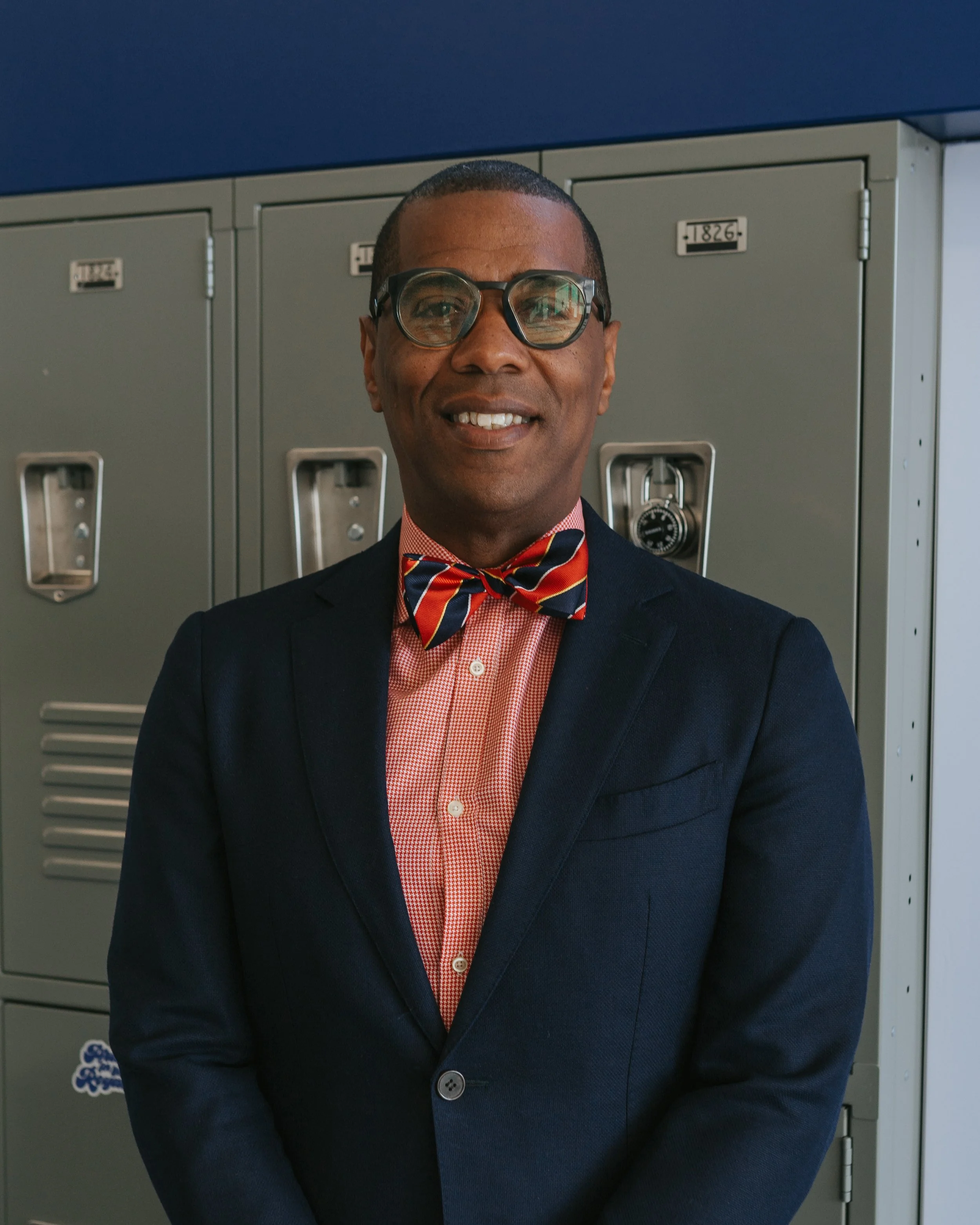 A man in a navy blazer, checkered red shirt, and striped bow tie stands in front of school lockers.