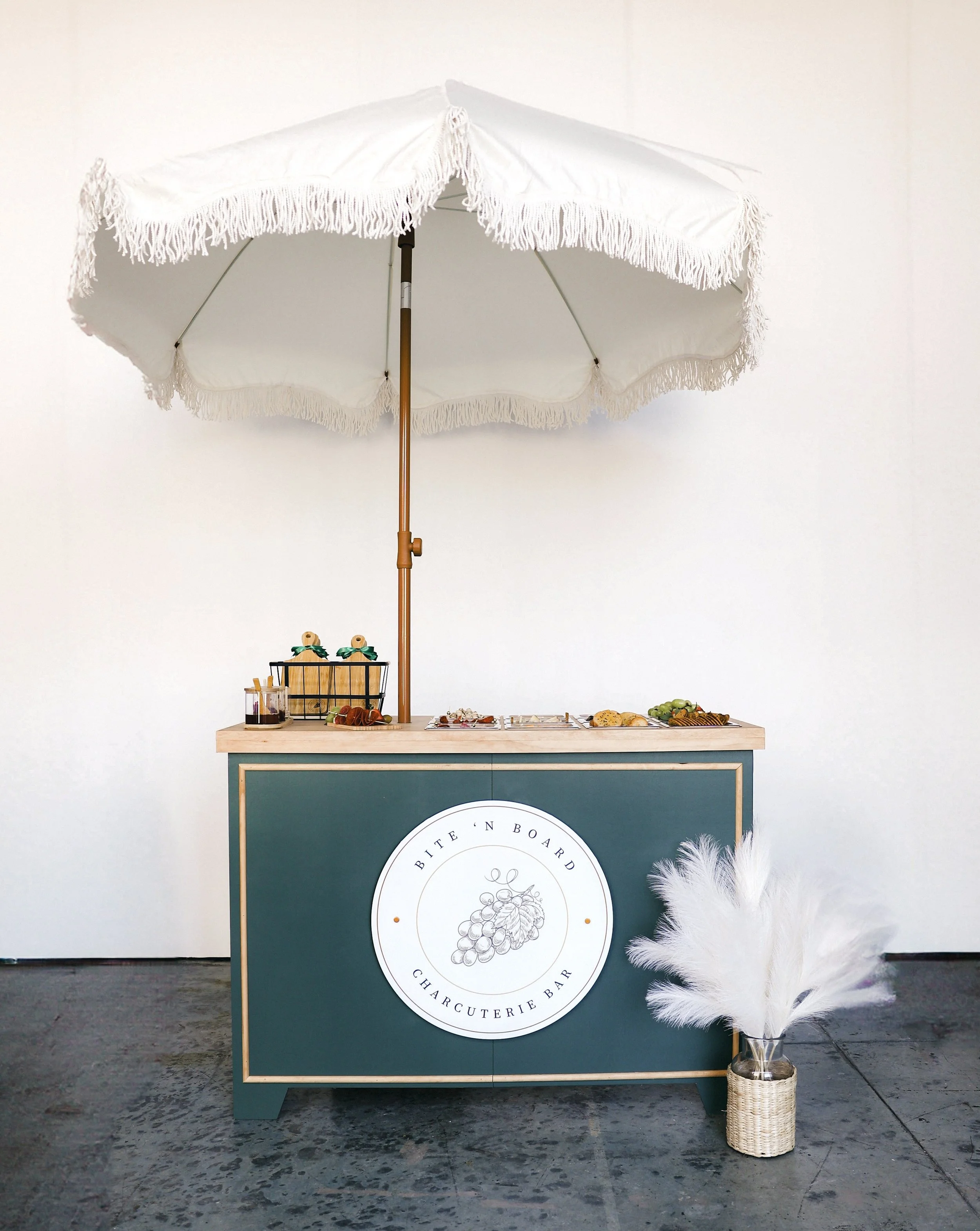 Charcuterie cart with a white umbrella, cart is green with brown accents.