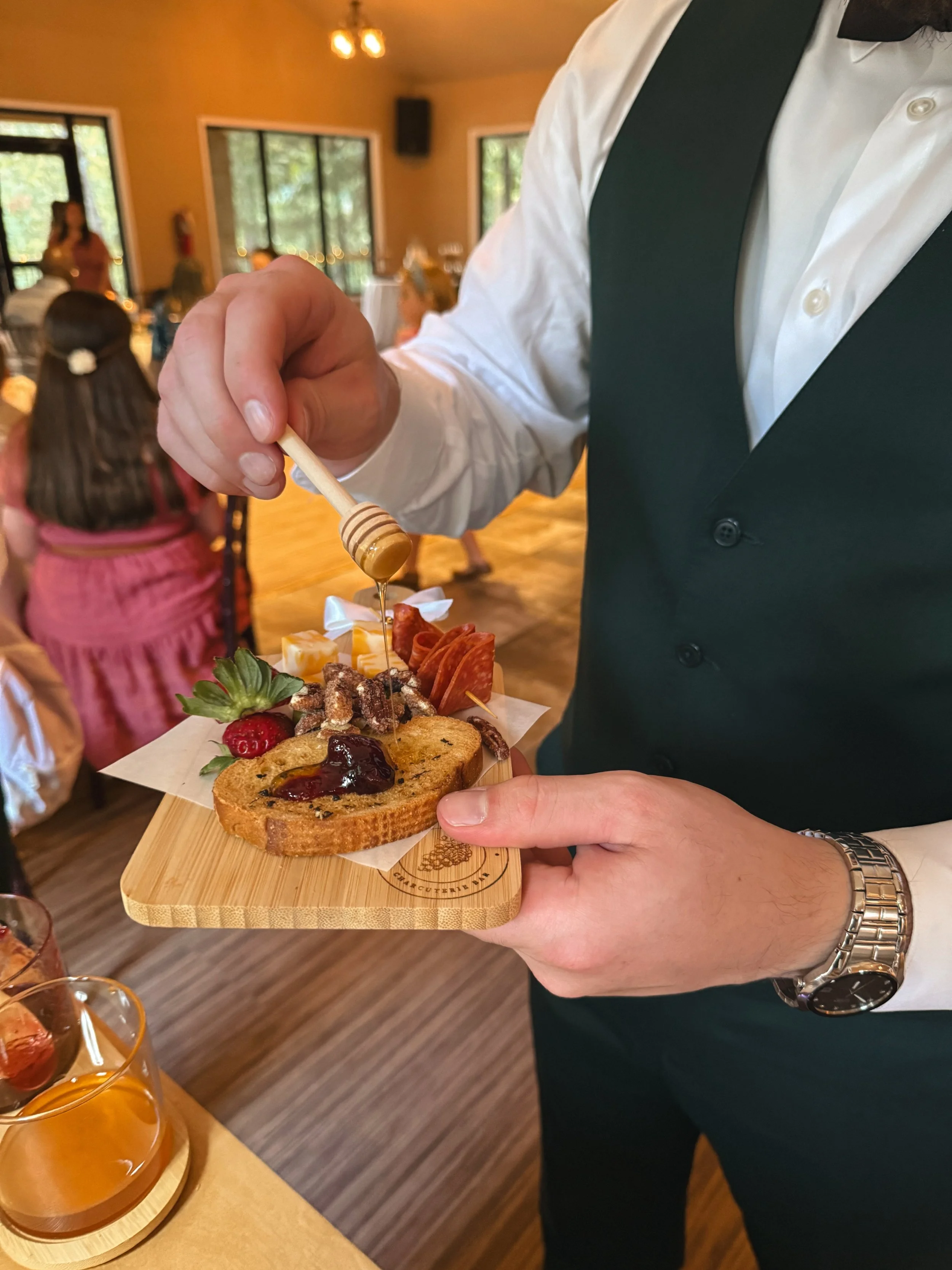 Honey dripping from a honey stirring stick onto food that is displayed on a mini charcuterie board with person holding it.