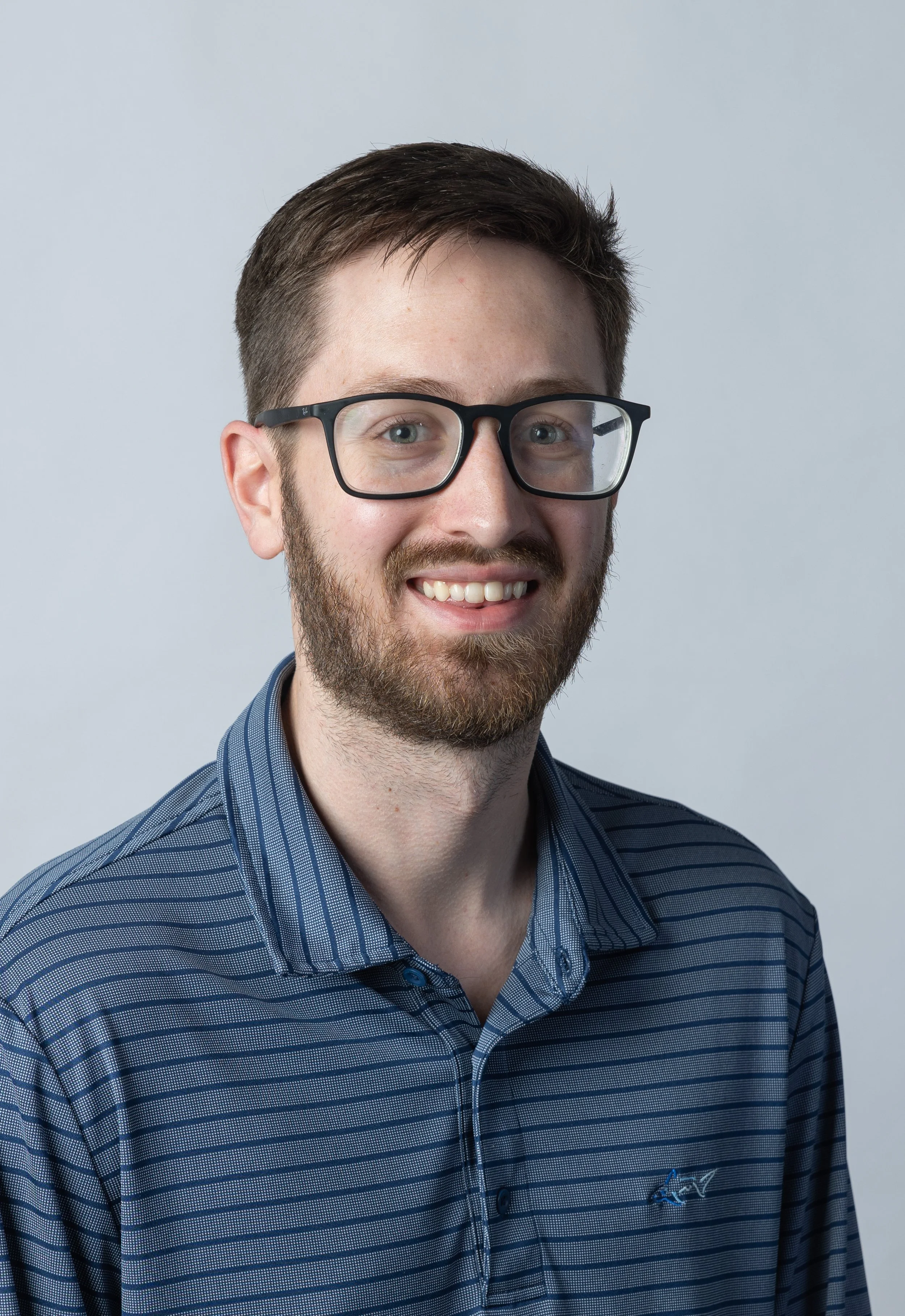 A young man with short brown hair, glasses, and a beard, wearing a blue striped shirt, smiling against a light gray background.