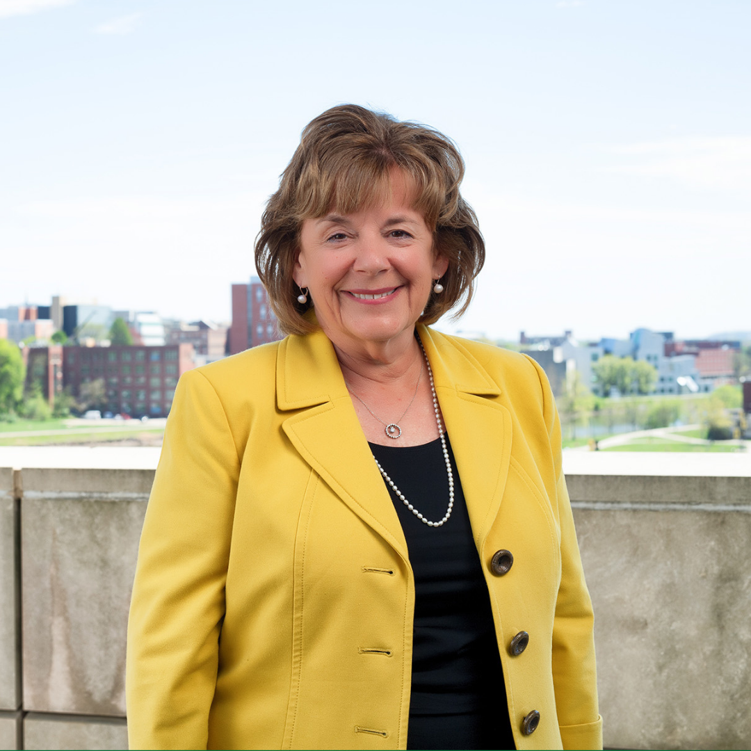 A smiling middle-aged woman with short brown hair wearing a yellow blazer, a black top, pearl earrings, and a pearl necklace. She is standing outdoors on a balcony with a cityscape and river in the background.