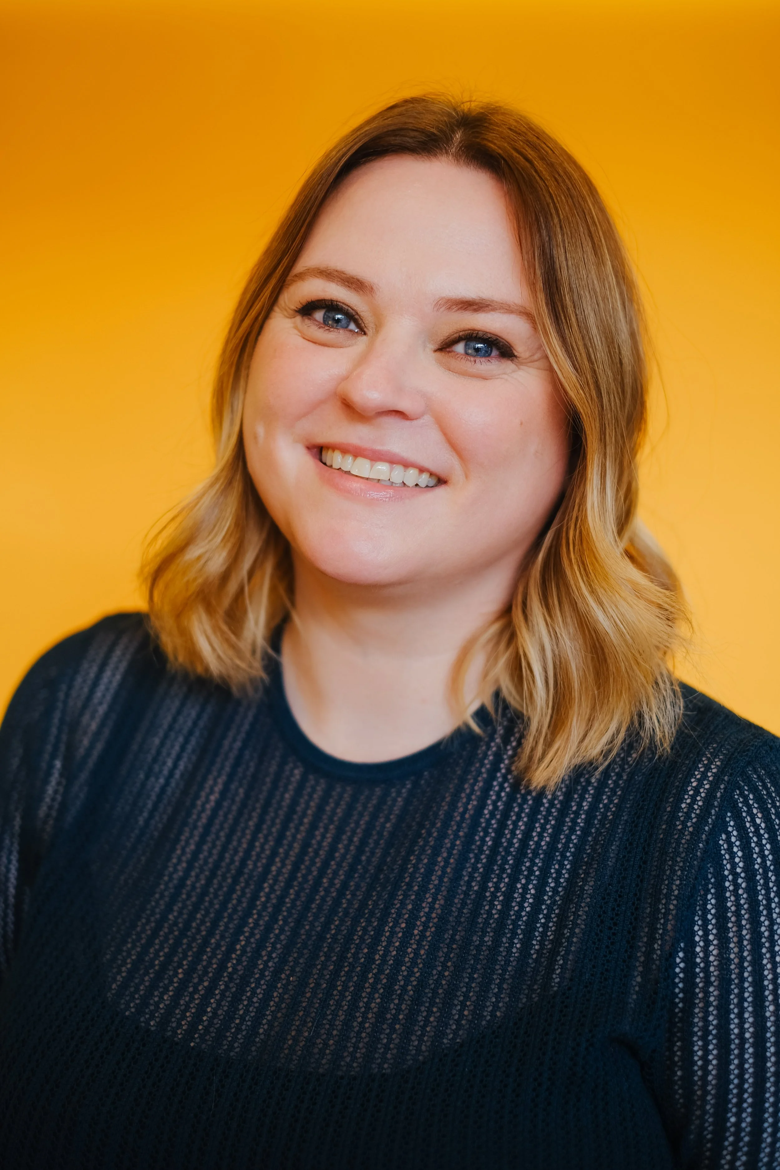 A smiling woman (Caroline) with shoulder-length wavy blonde hair and blue eyes, wearing a black, striped, semi-sheer top, standing against a solid yellow background.