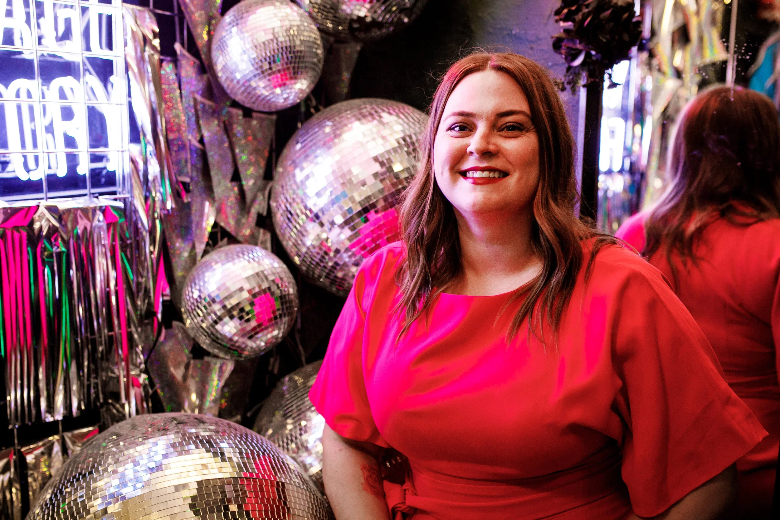 A young woman (Caroline) with long brown hair, smiling in front of a wall of disco balls and shiny metallic decorations, wearing a bright pink dress.