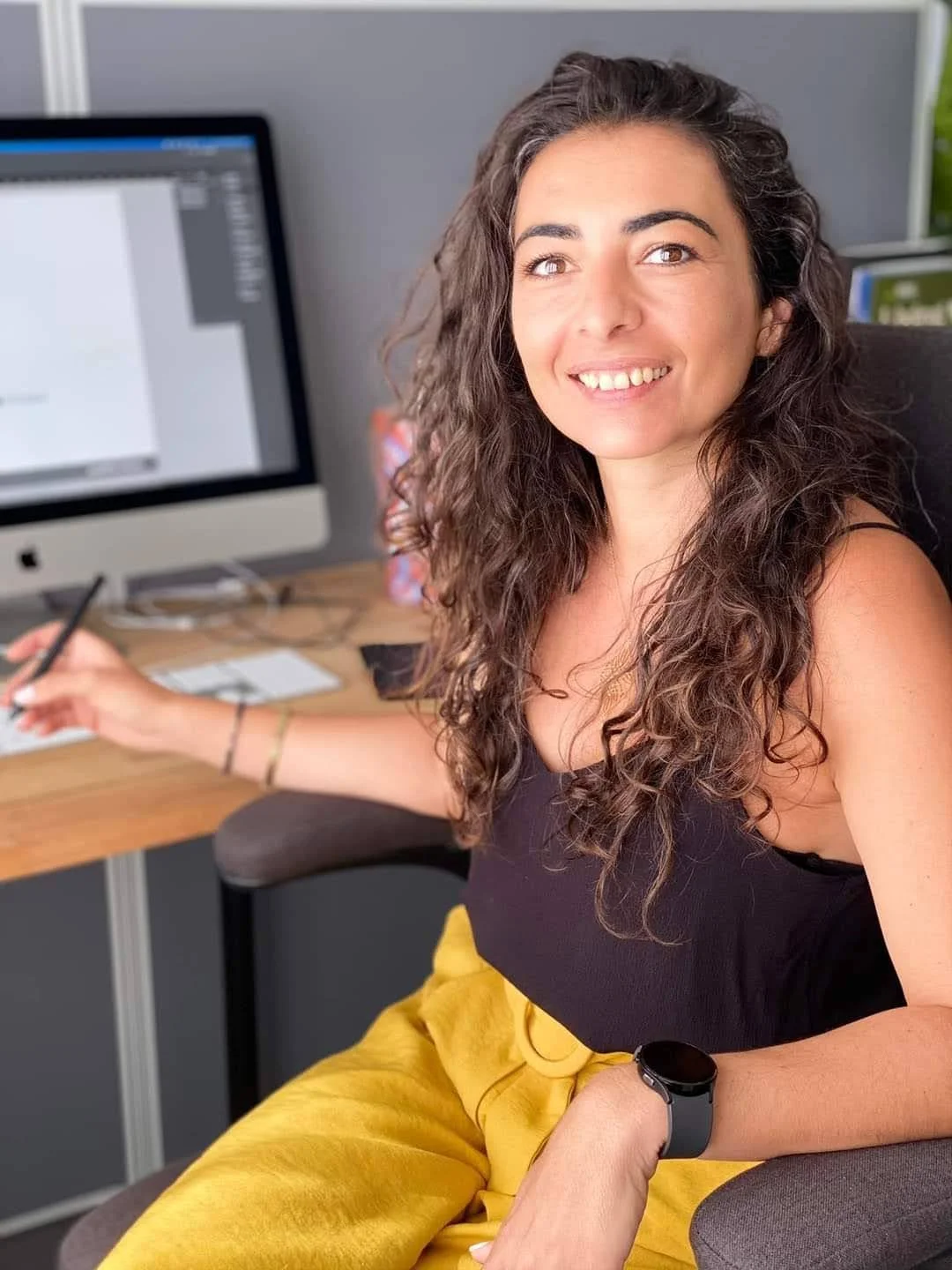 A woman with long curly brown hair smiling while sitting at a desk with a computer, wearing a black top, yellow pants, and a black smartwatch.
