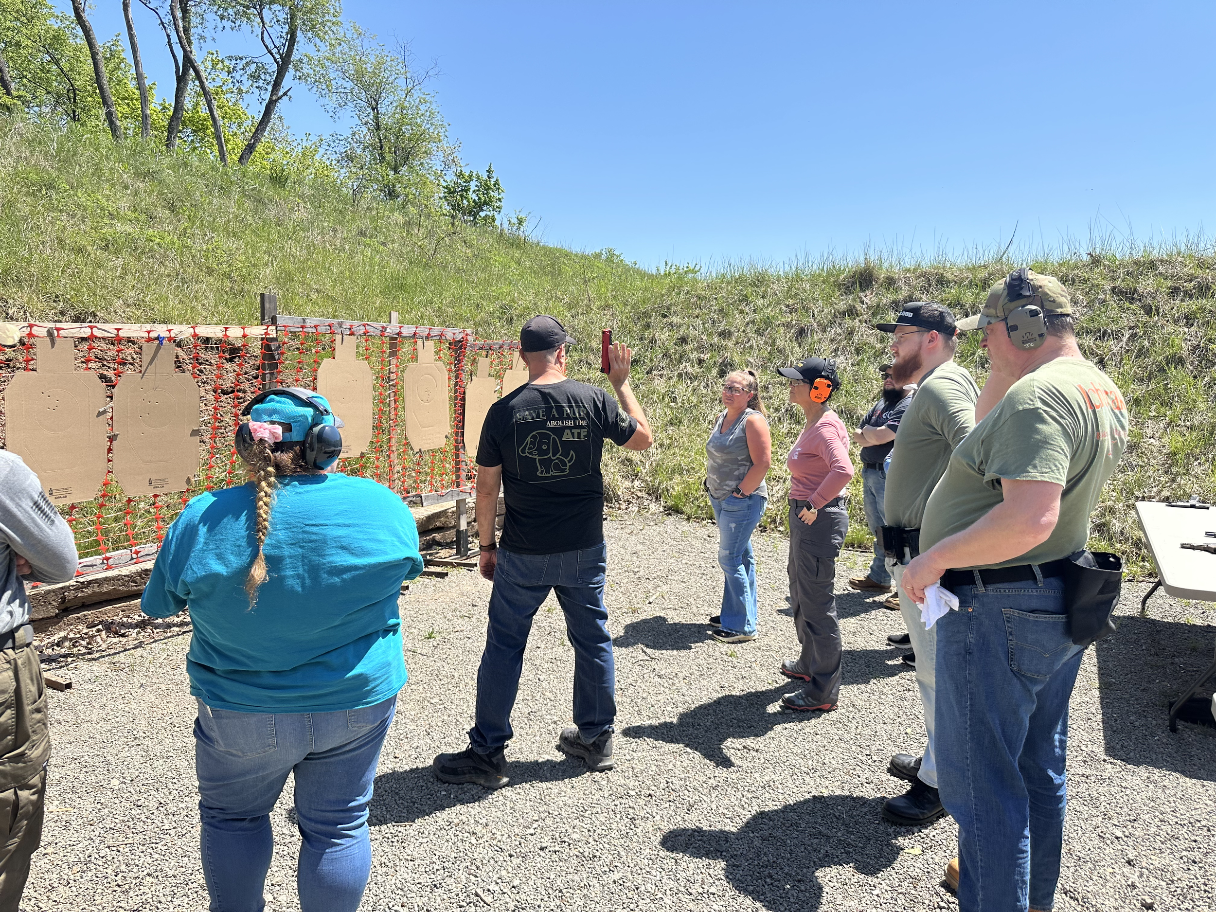 A group of people at a shooting range, with some wearing eye and ear protection, standing in line and facing targets on a backstop, outdoors on a sunny day.