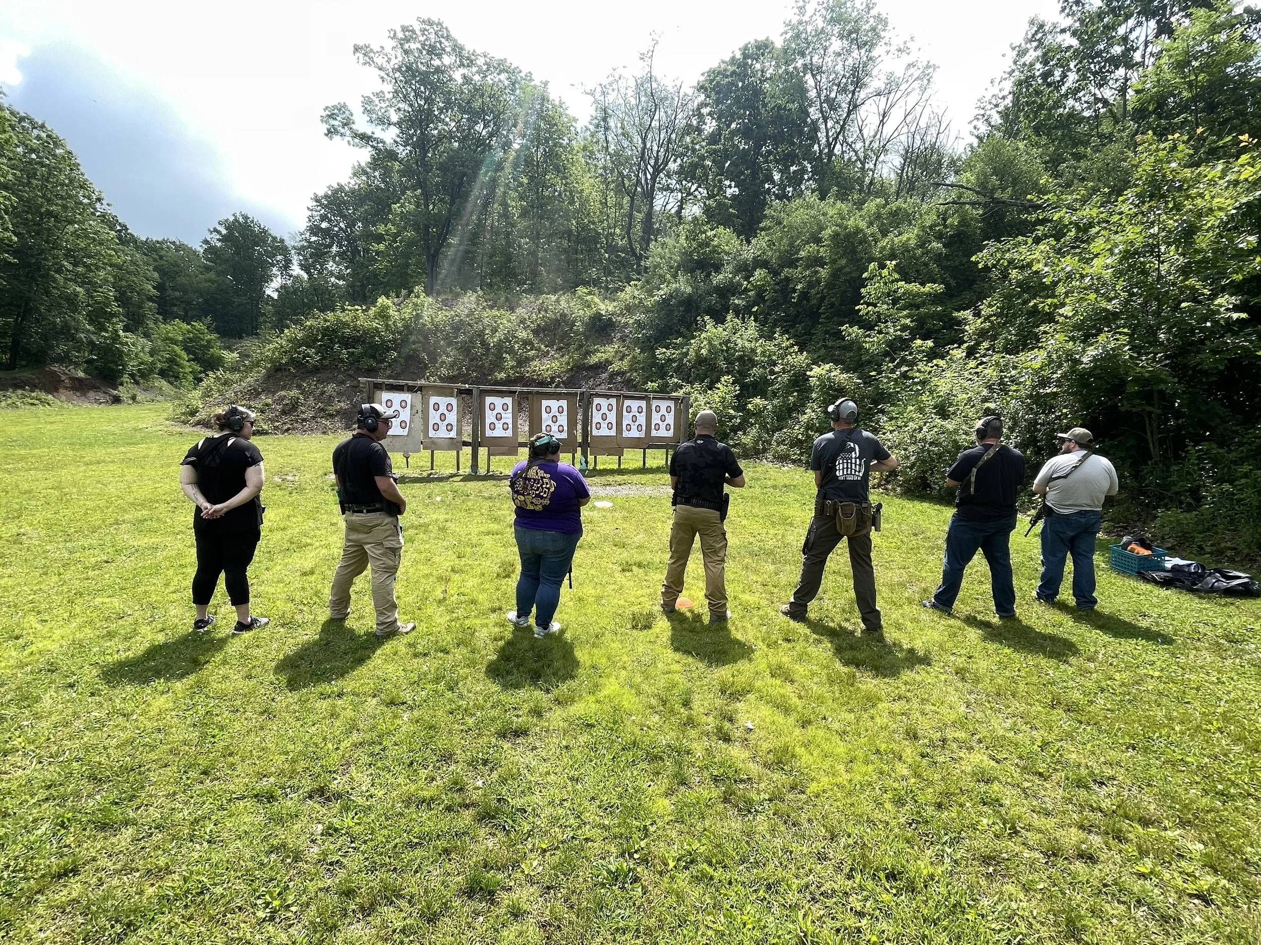 A group of people practicing shooting at outdoor target range in a grassy area with trees in the background.