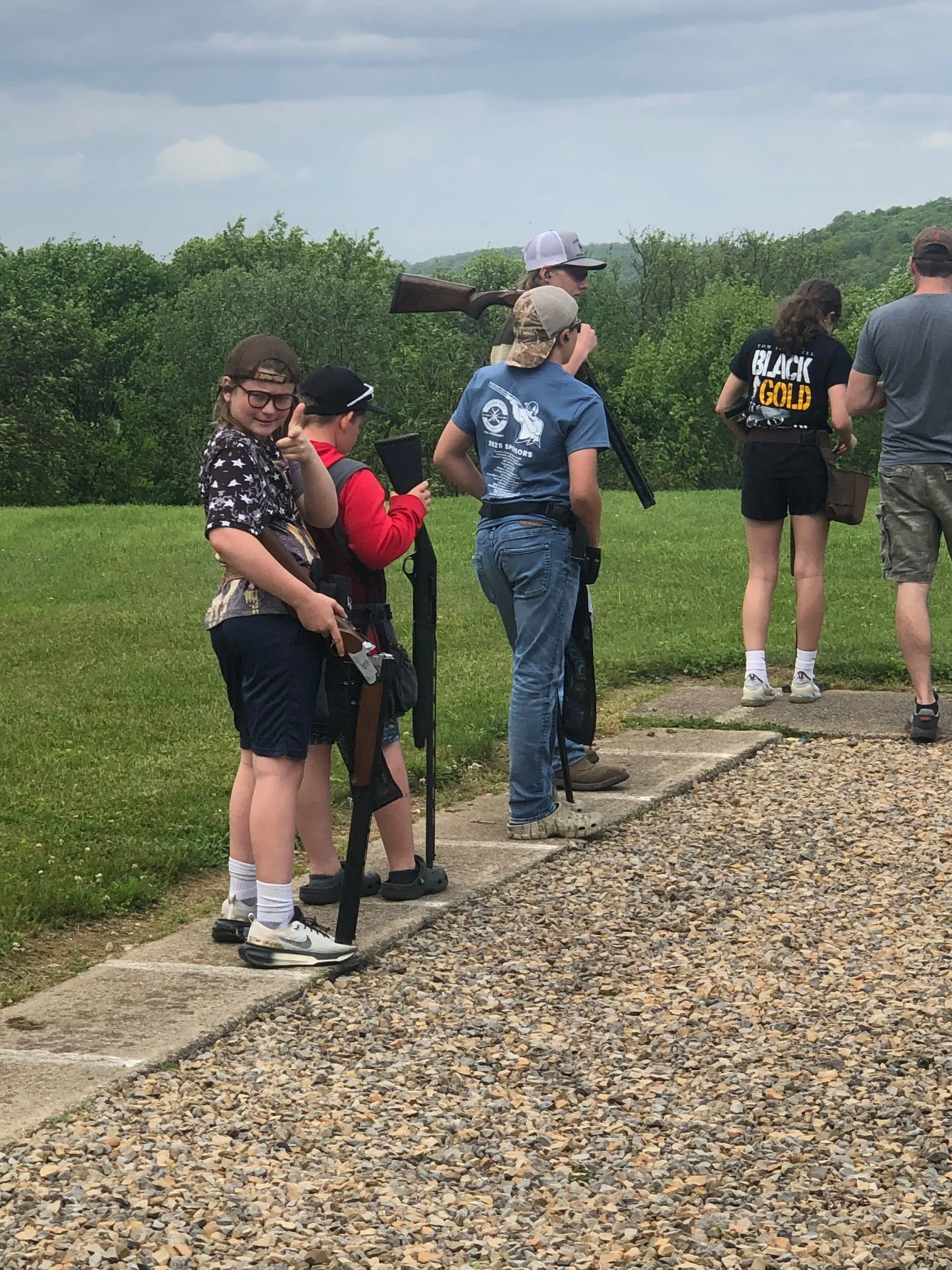 Group of children and adults at an outdoor shooting range, some holding rifles, on a cloudy day with green trees in the background.