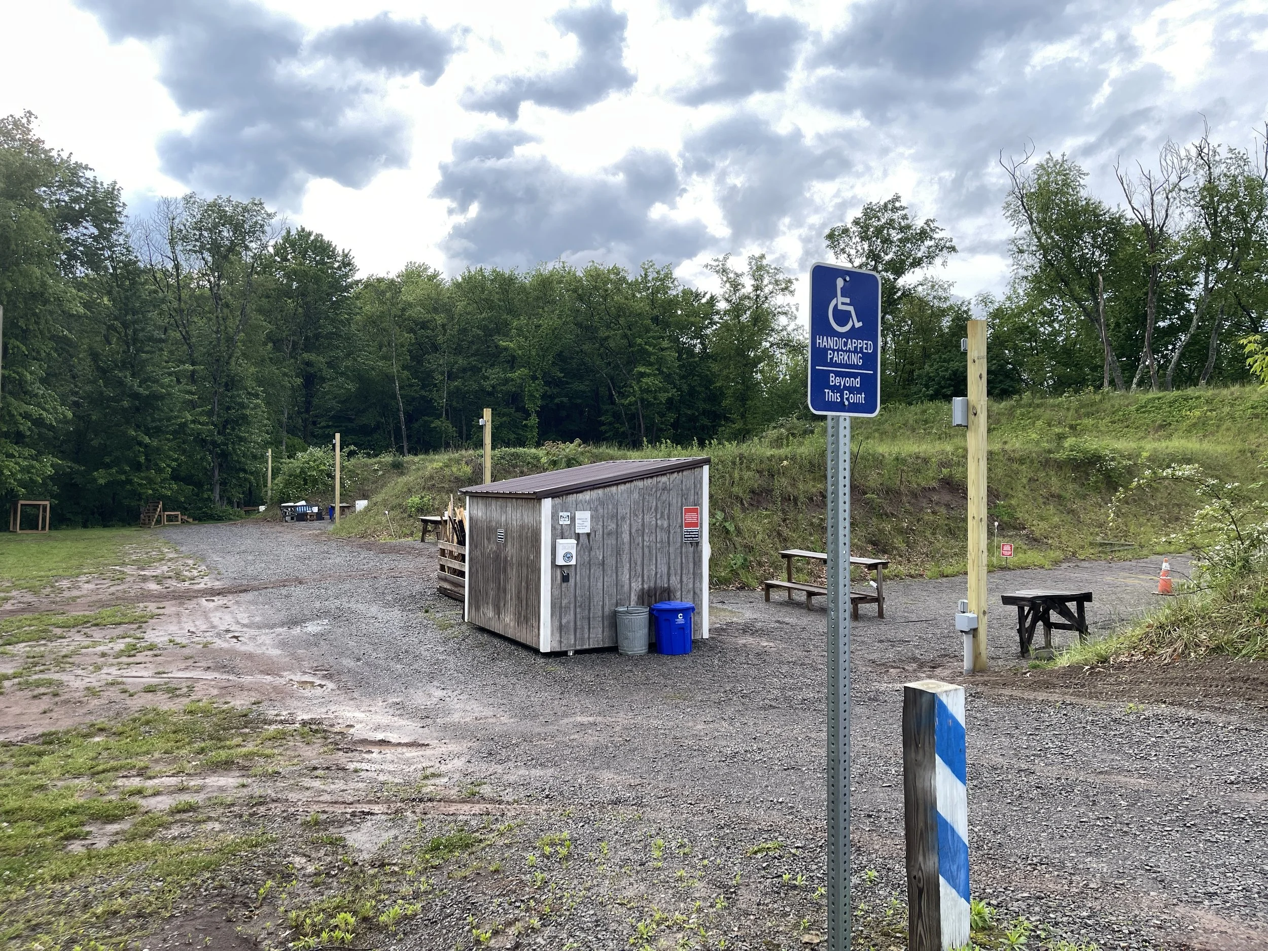 A gravel parking area adjacent to a wooded hill with a blue sign indicating handicapped parking beyond this point. There are picnic tables, a small wooden shed, trash cans, and a wooden post with a white and blue striped marker. The background featur